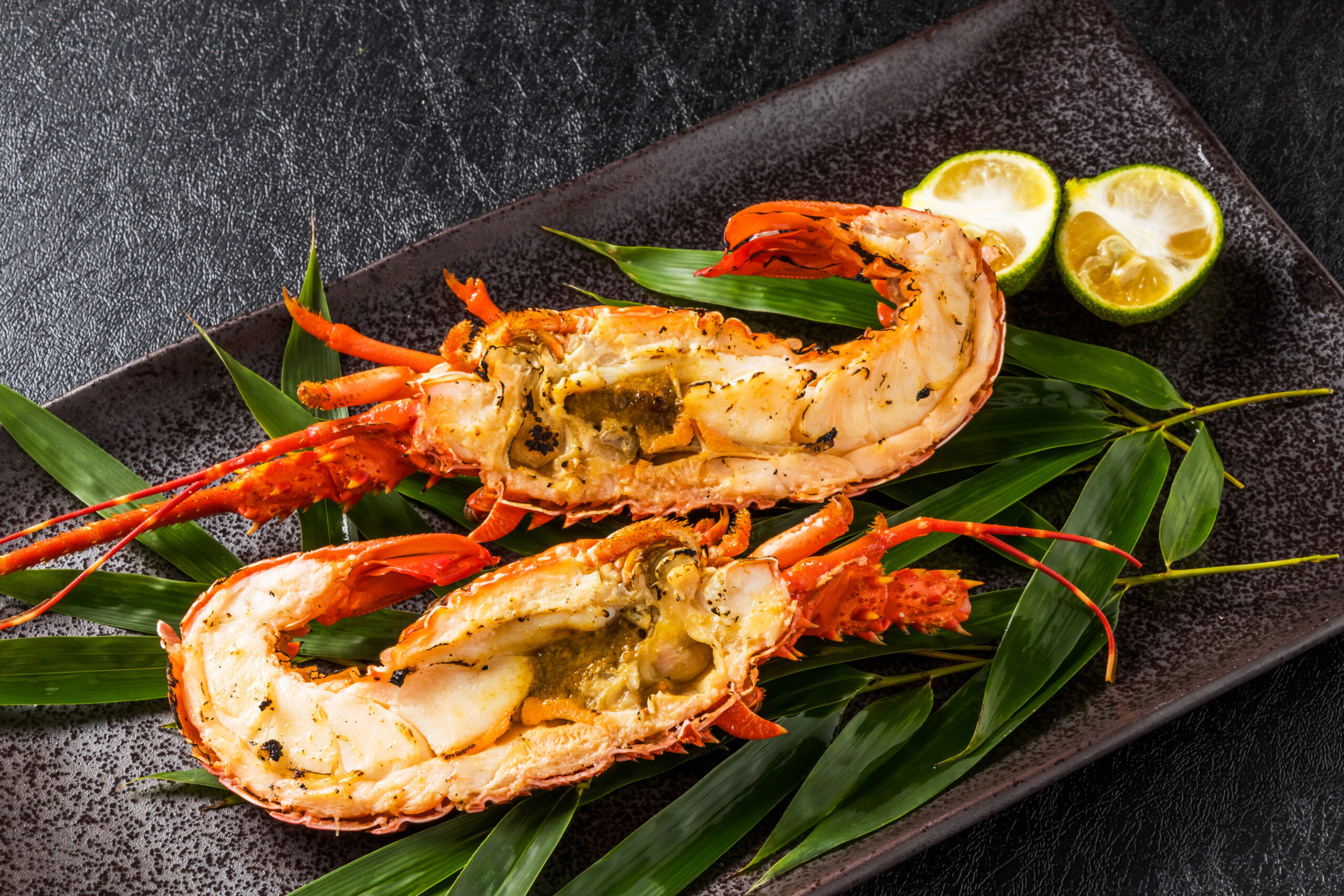 Grilled lobster tails split open on a dark plate with lime wedges and green leaves as garnish.