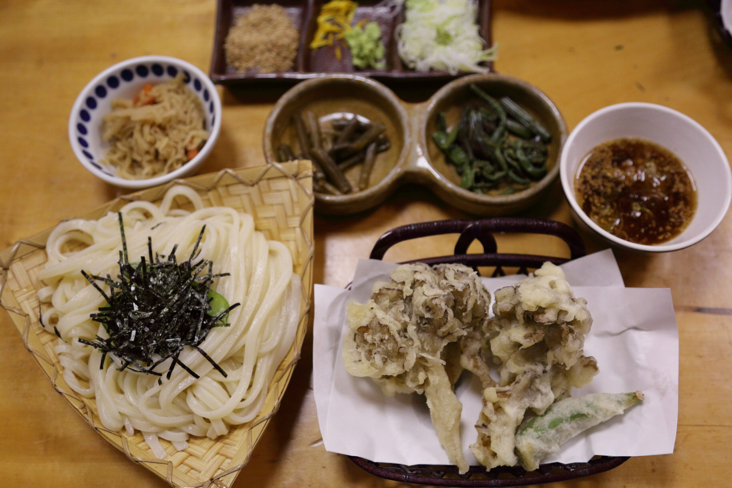 Thick udon noodles with shredded seaweed in a woven basket, plus assorted tempura and a dipping sauce on the side.