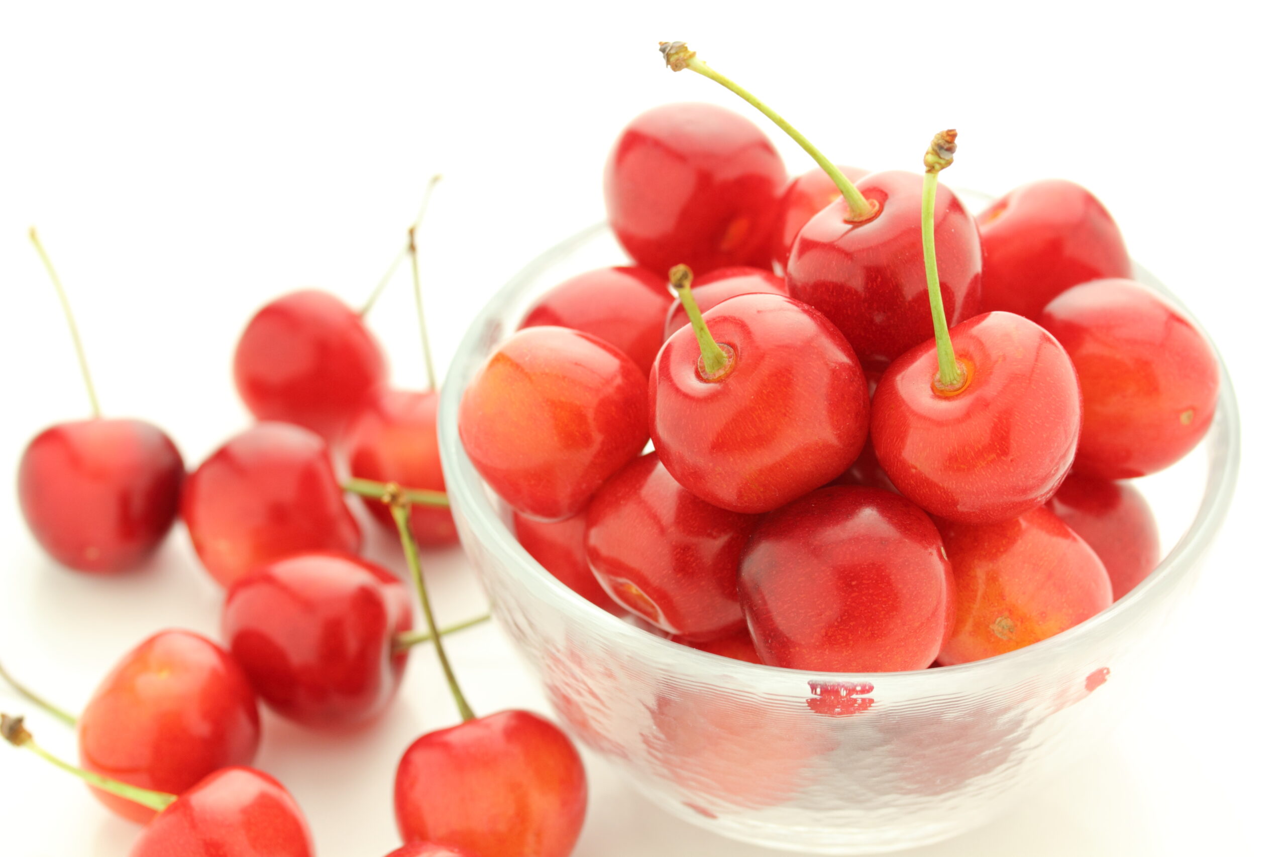 Pile of bright red cherries with green stems in a clear glass bowl against a white background, close-up.