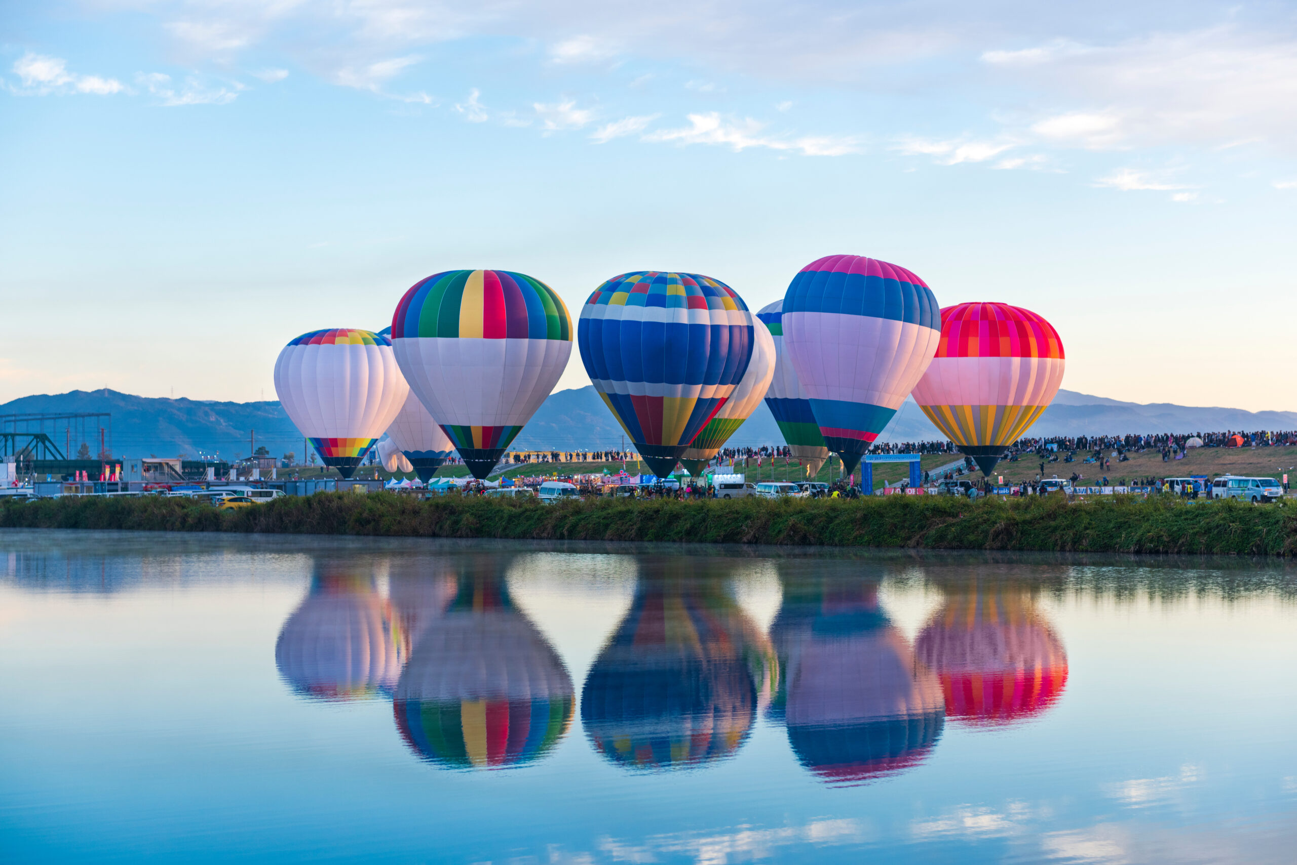 Colorful hot air balloons ready for flight on a grassy field beside a calm lake, with a crowd in the distance and distant hills under a blue sky, mirrored in the water.