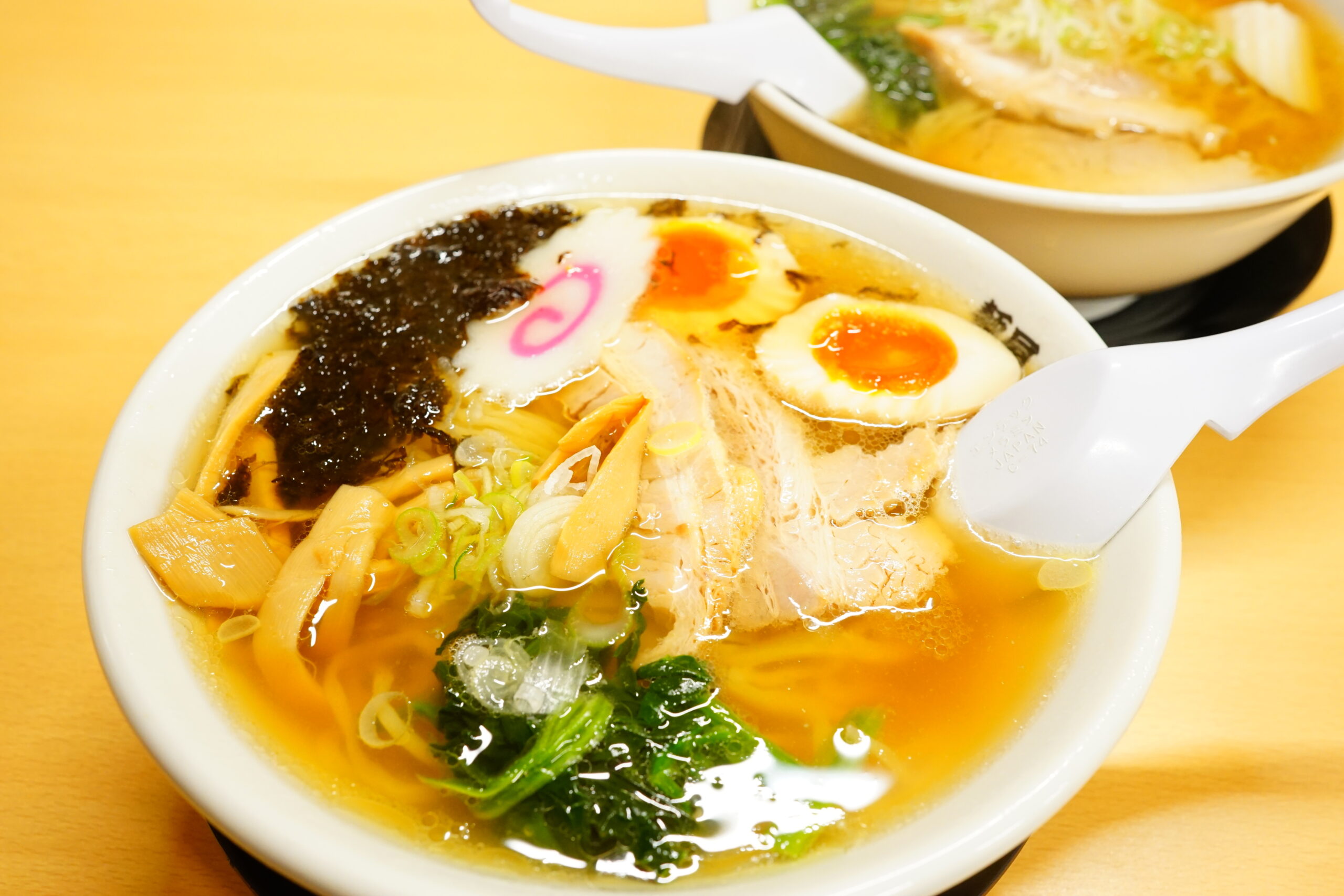 Bowl of ramen with soft-boiled egg, seaweed, green onions, bamboo shoots, and narutomaki in a rich broth; another bowl visible in the background