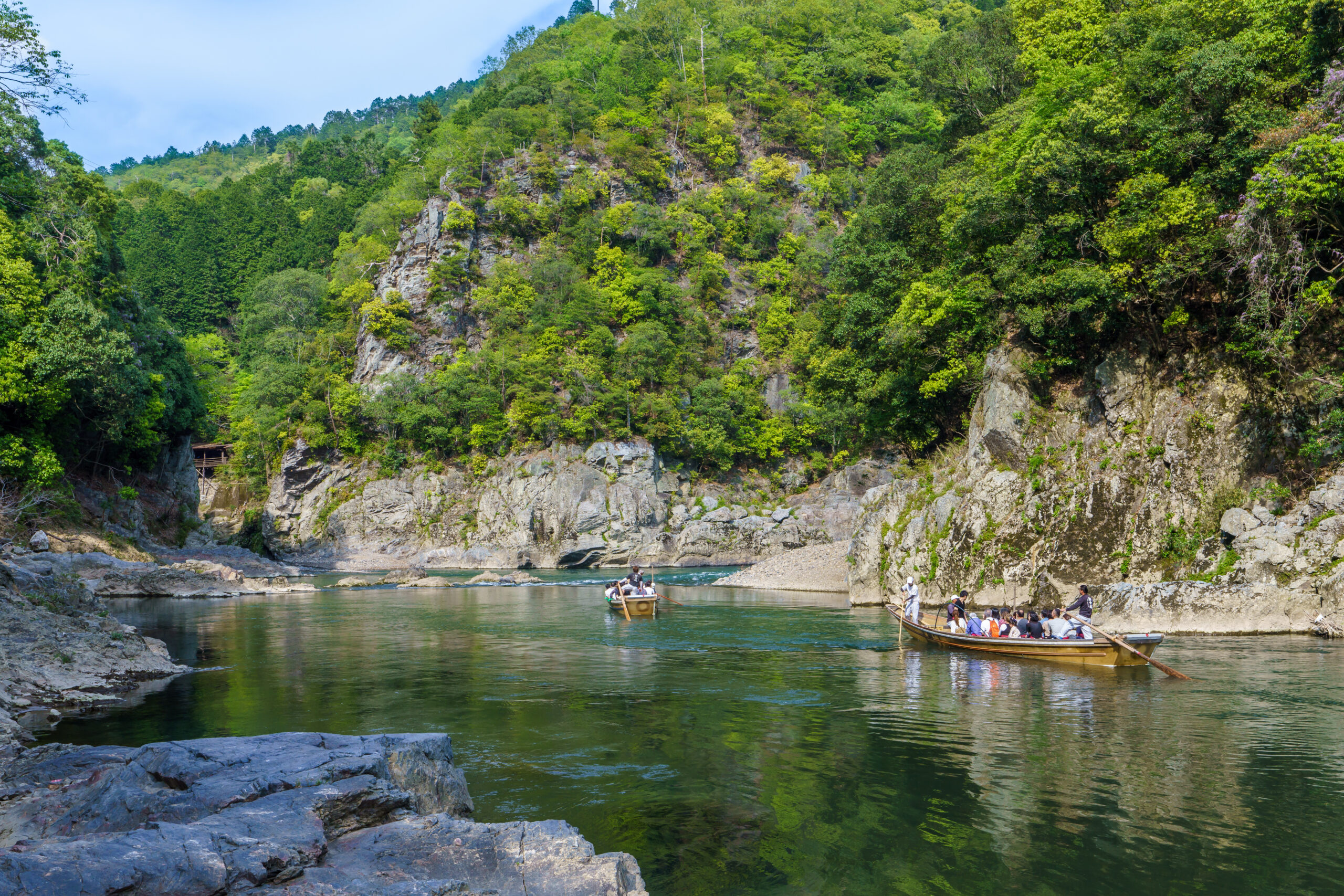 Group of people in a boat navigating a calm river flanked by rocky cliffs and dense green forested hillsides.