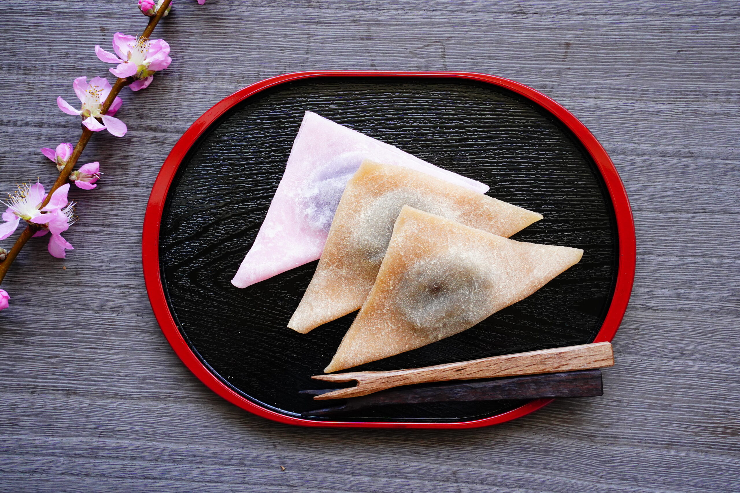 Assorted pastel folded mochi triangles on a black plate with a red rim, beside a pink blossom branch on a wooden surface.