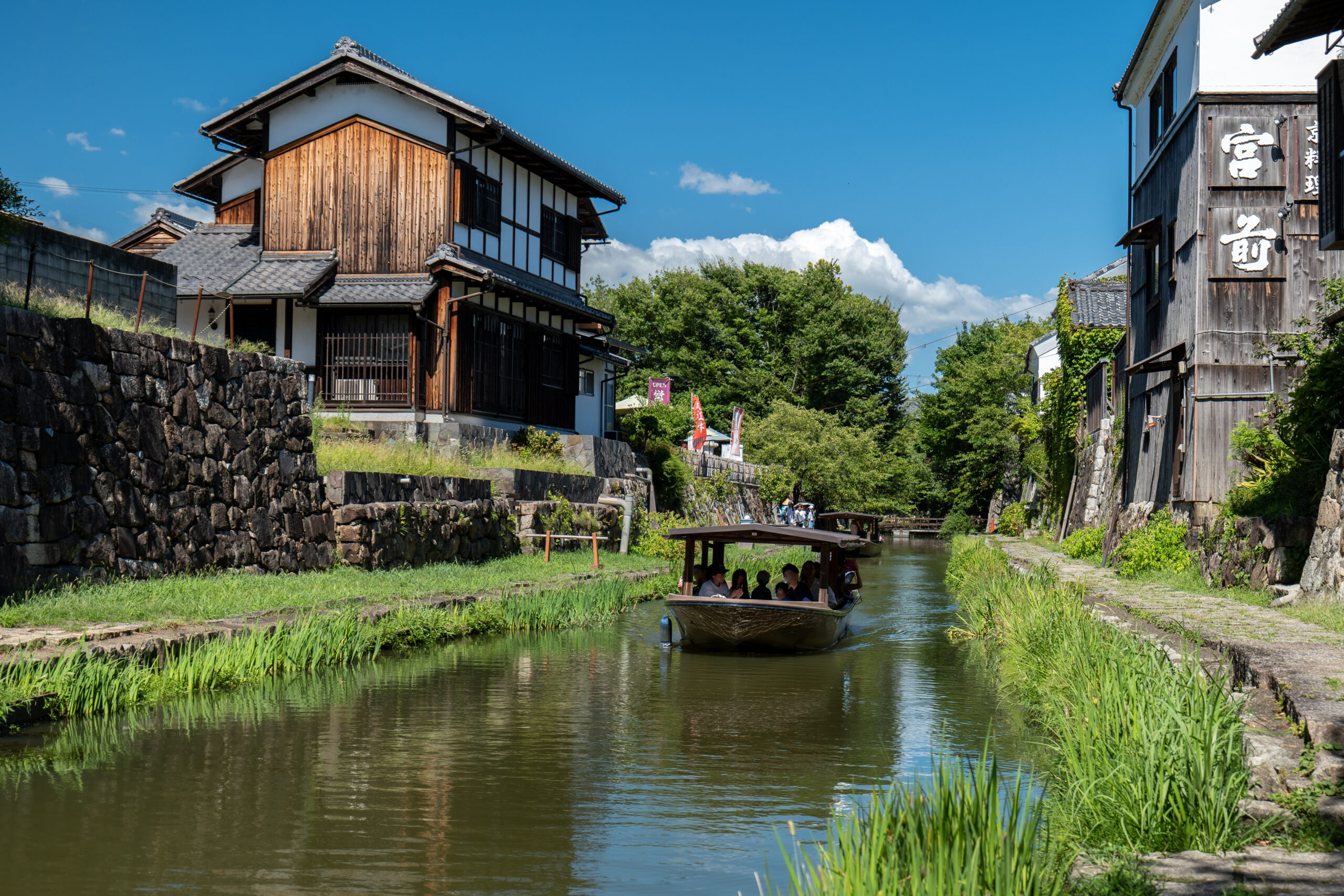 Passenger boat gliding along a narrow canal bordered by traditional wooden buildings and stone walls under a blue sky.