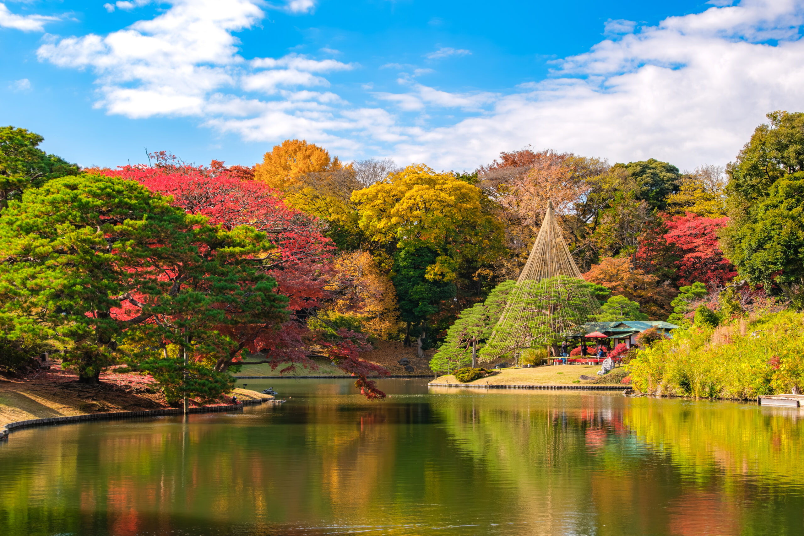Autumn trees with red, yellow, and green foliage beside a lake under a blue sky; a conical wooden structure is visible among the trees in the distance.