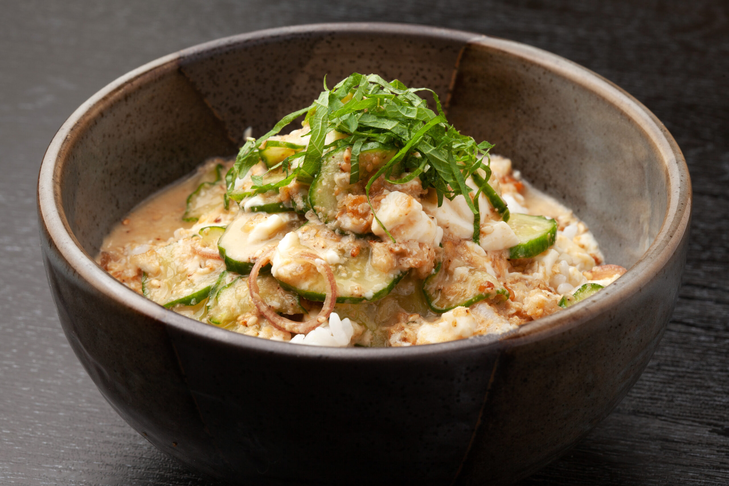 Clay bowl with a cucumber-based salad: sliced cucumbers, fresh herbs, and a creamy dressing over rice-filled bottomed dish.