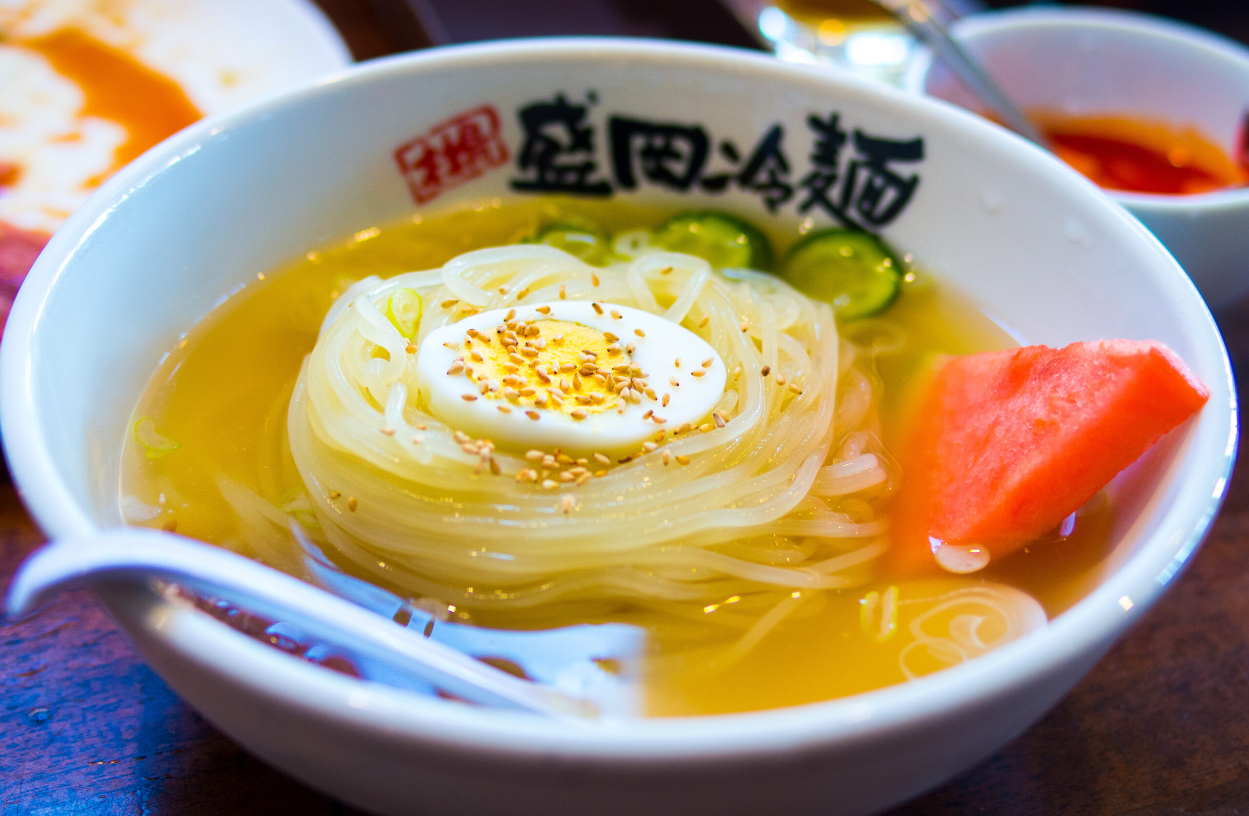 Bowl of noodle soup with a halved boiled egg sprinkled with sesame seeds, cucumber slices, and a watermelon wedge in broth.