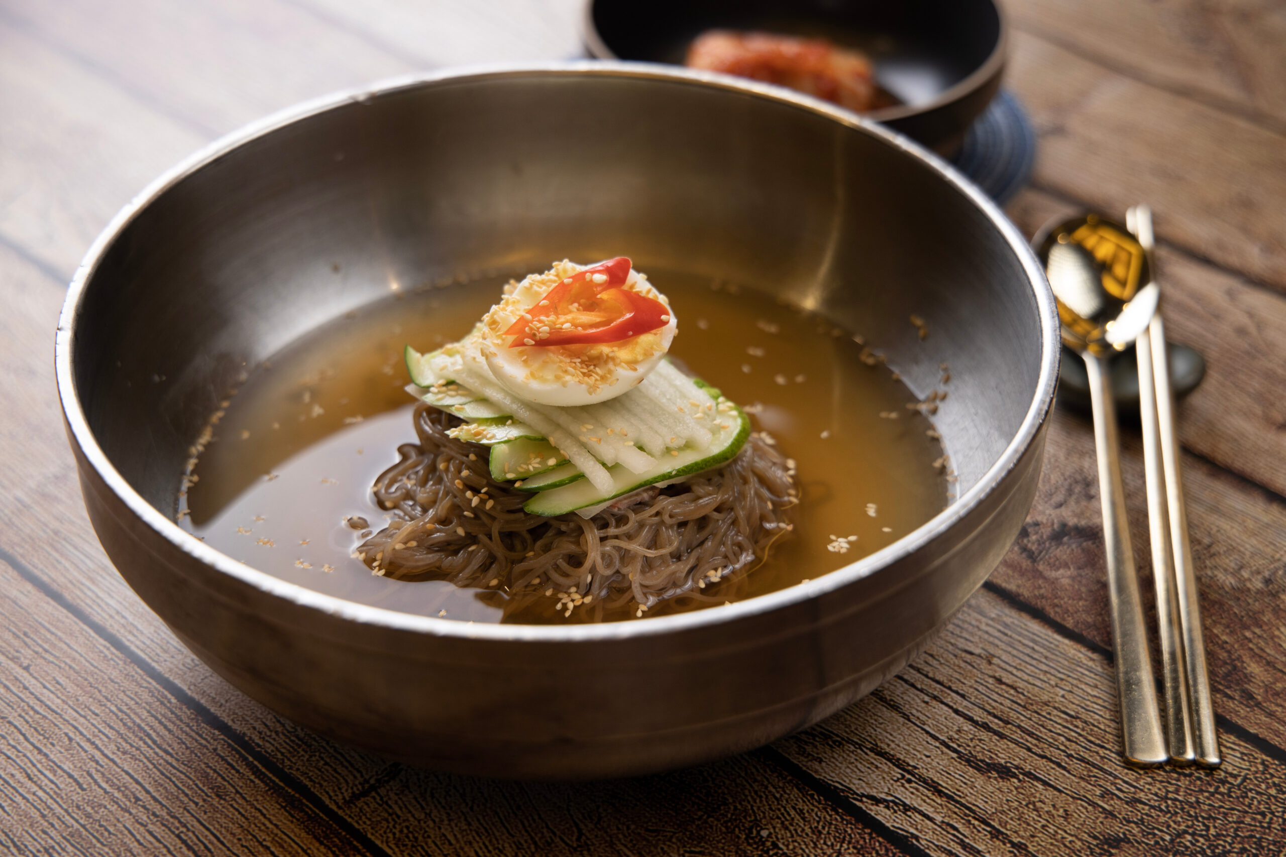 Metal bowl of noodle soup with brown broth and toppings: cucumber ribbons, sliced boiled egg, sesame seeds on top, served on a wooden table.