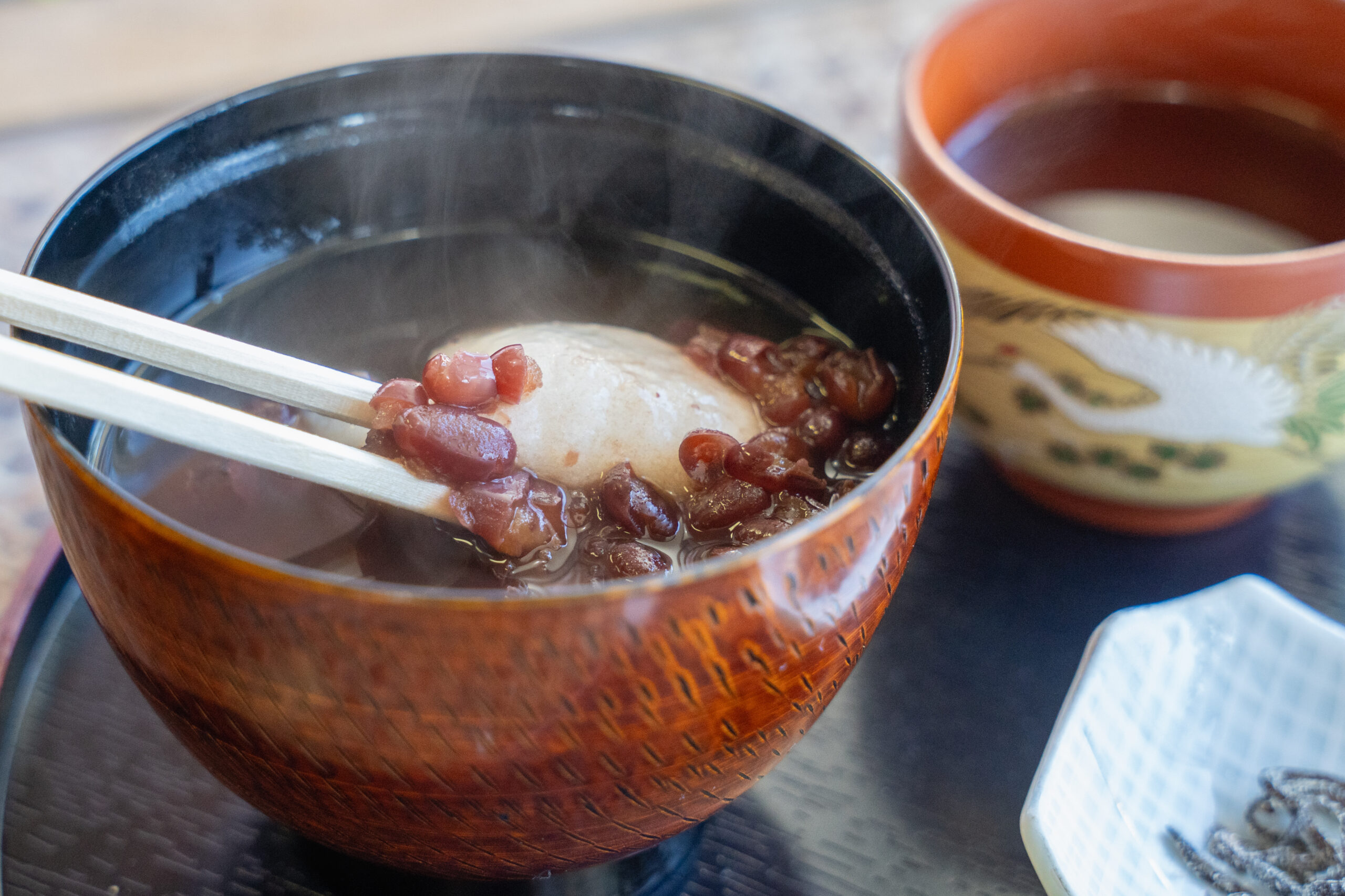 Lacquered orange bowl with hot red bean soup and a mochi ball, chopsticks resting on the rim.