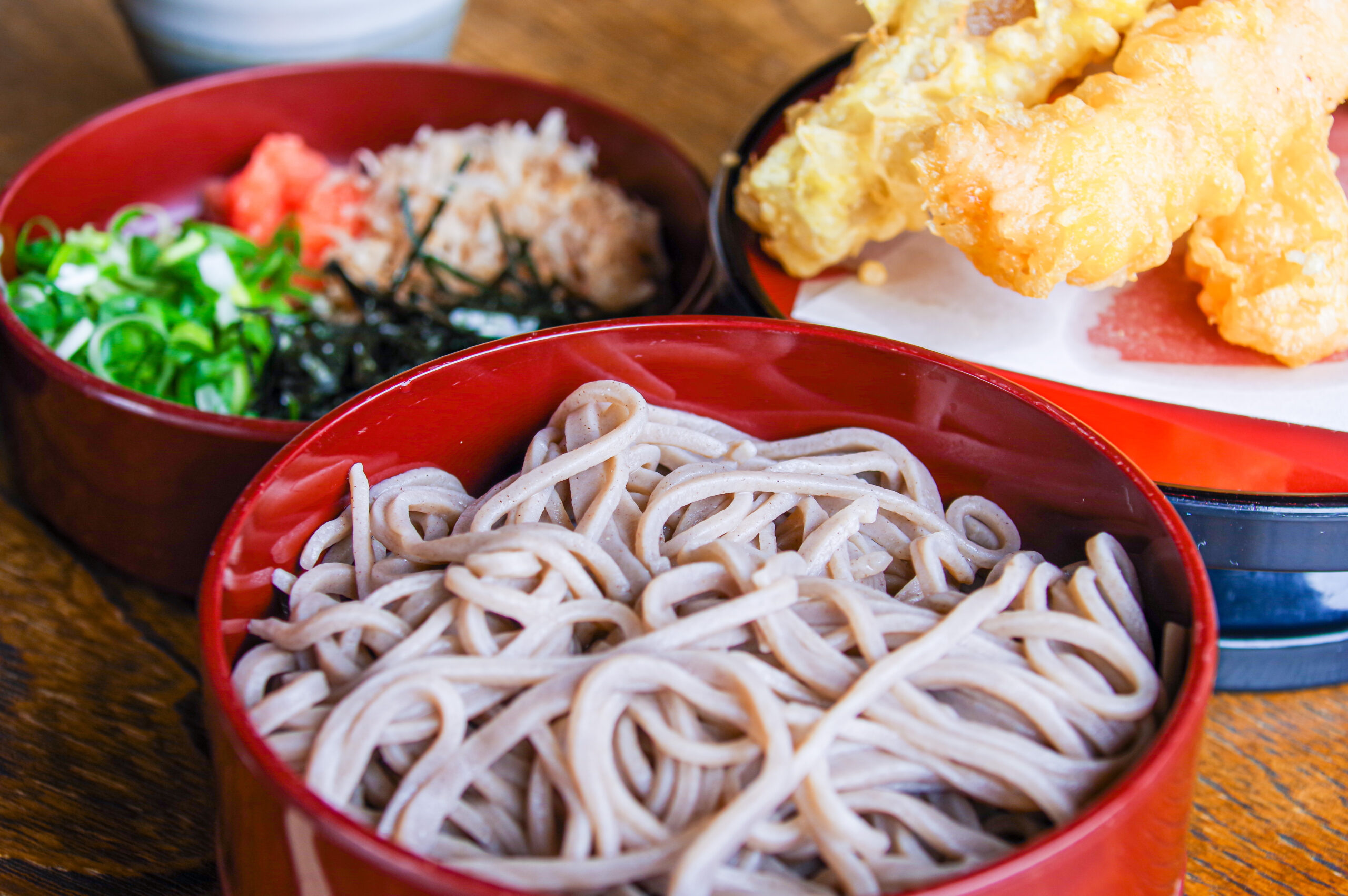 Red bowls of soba noodles with tempura and green onions on a wooden table, garnished with seaweed.
