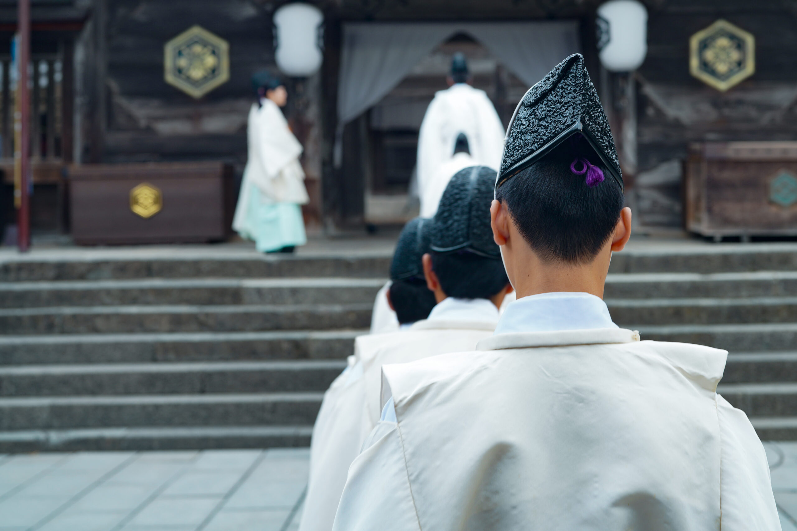 Back view of a person in white ceremonial robes and a black patterned hat, facing a temple altar as others in similar attire stand on stone steps.