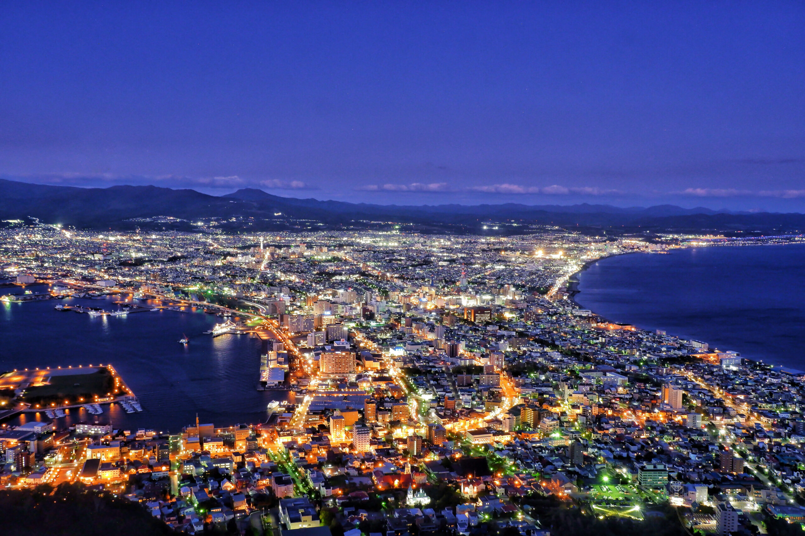 Nighttime aerial view of a coastal city illuminated by street and building lights, with mountains in the distance and a dark blue sky above.