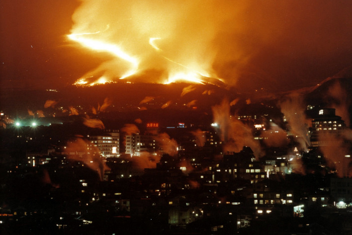Nighttime city below a volcano erupting with lava glow and billowing smoke in the distance.