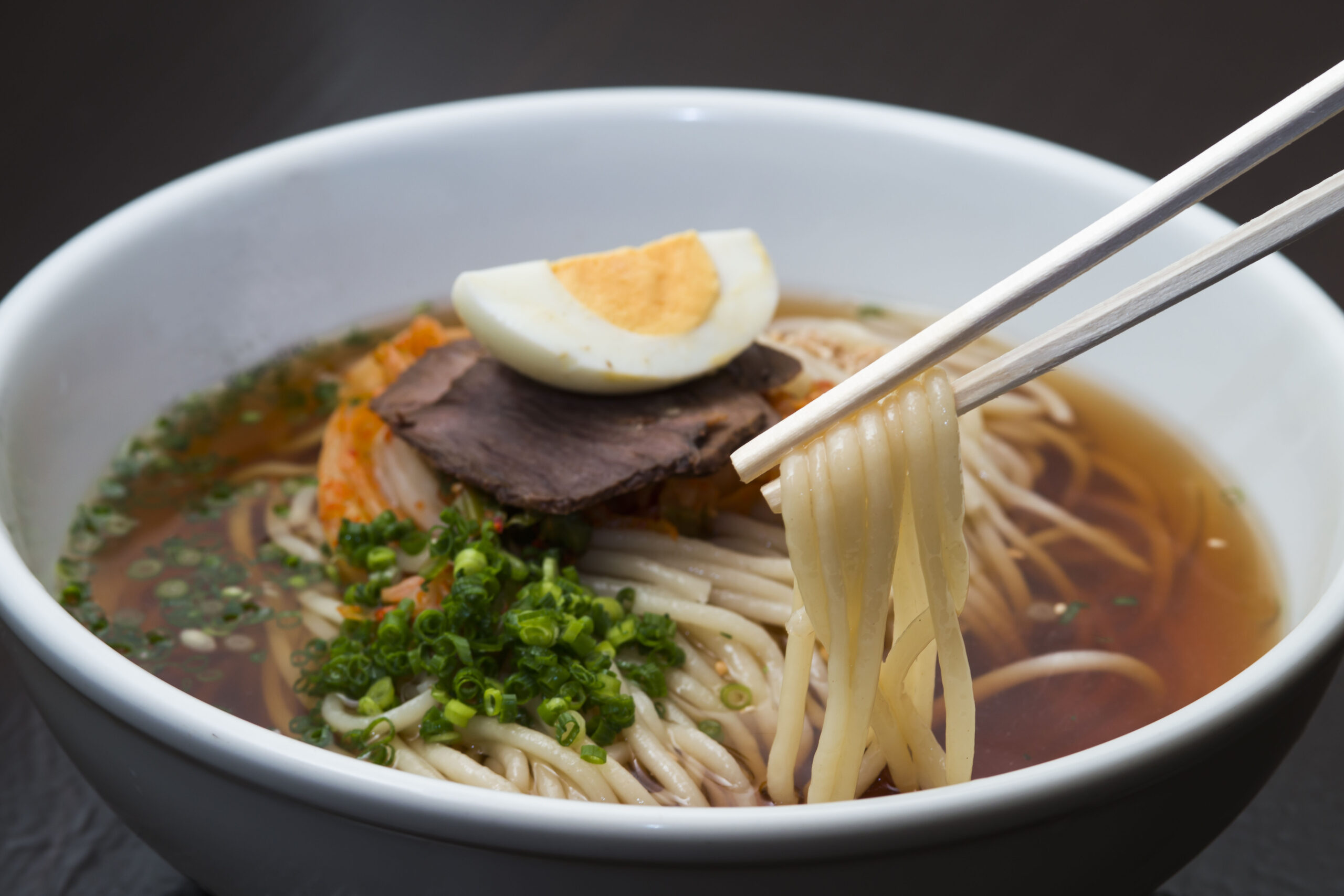 Bowl of ramen with sliced beef, half a boiled egg, green onions, and chopsticks picking up noodles