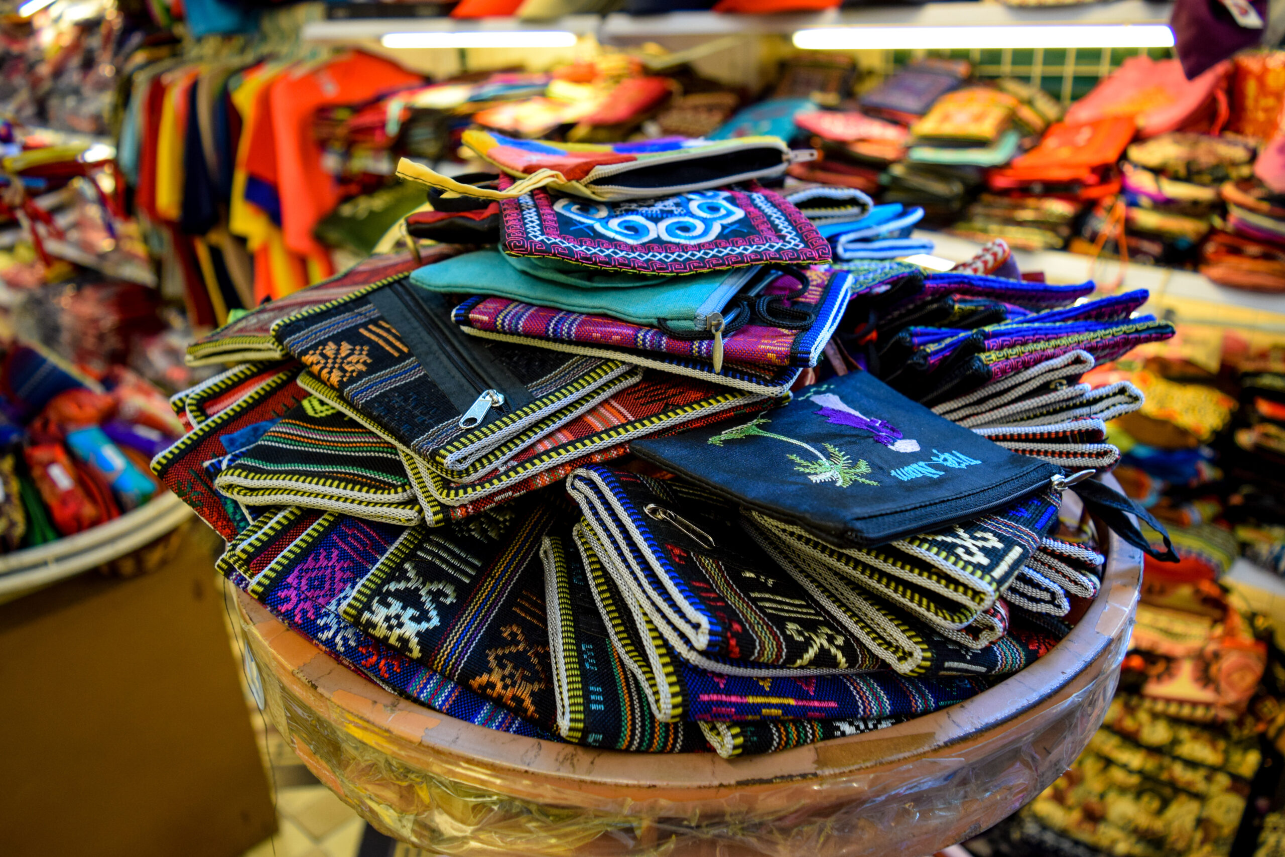 Pile of colorful embroidered fabric pouches with zippers stacked on a basket at a market stall.