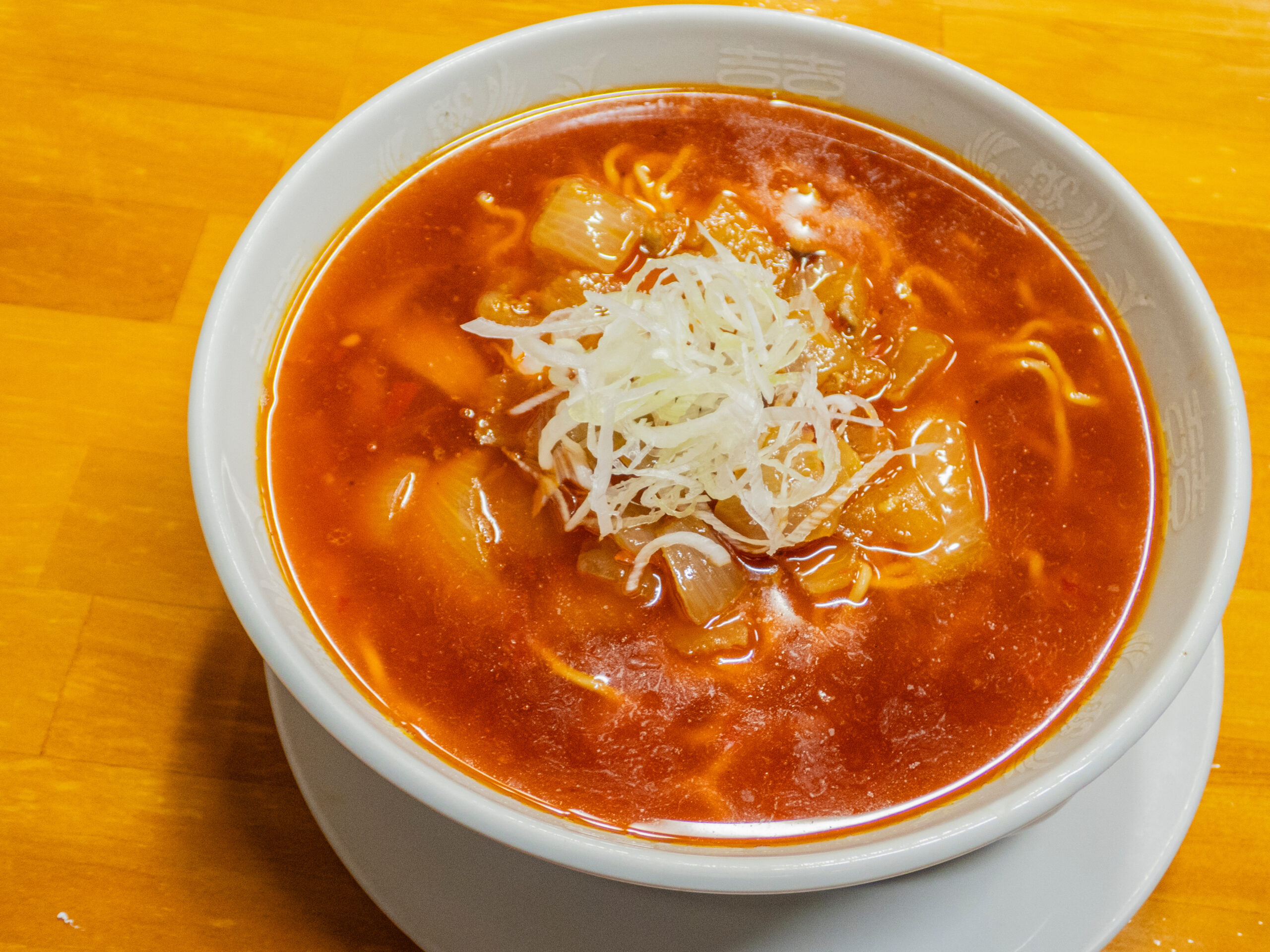 Bowl of red tomato-based soup with noodles and shredded cheese on top, served on a wooden table.