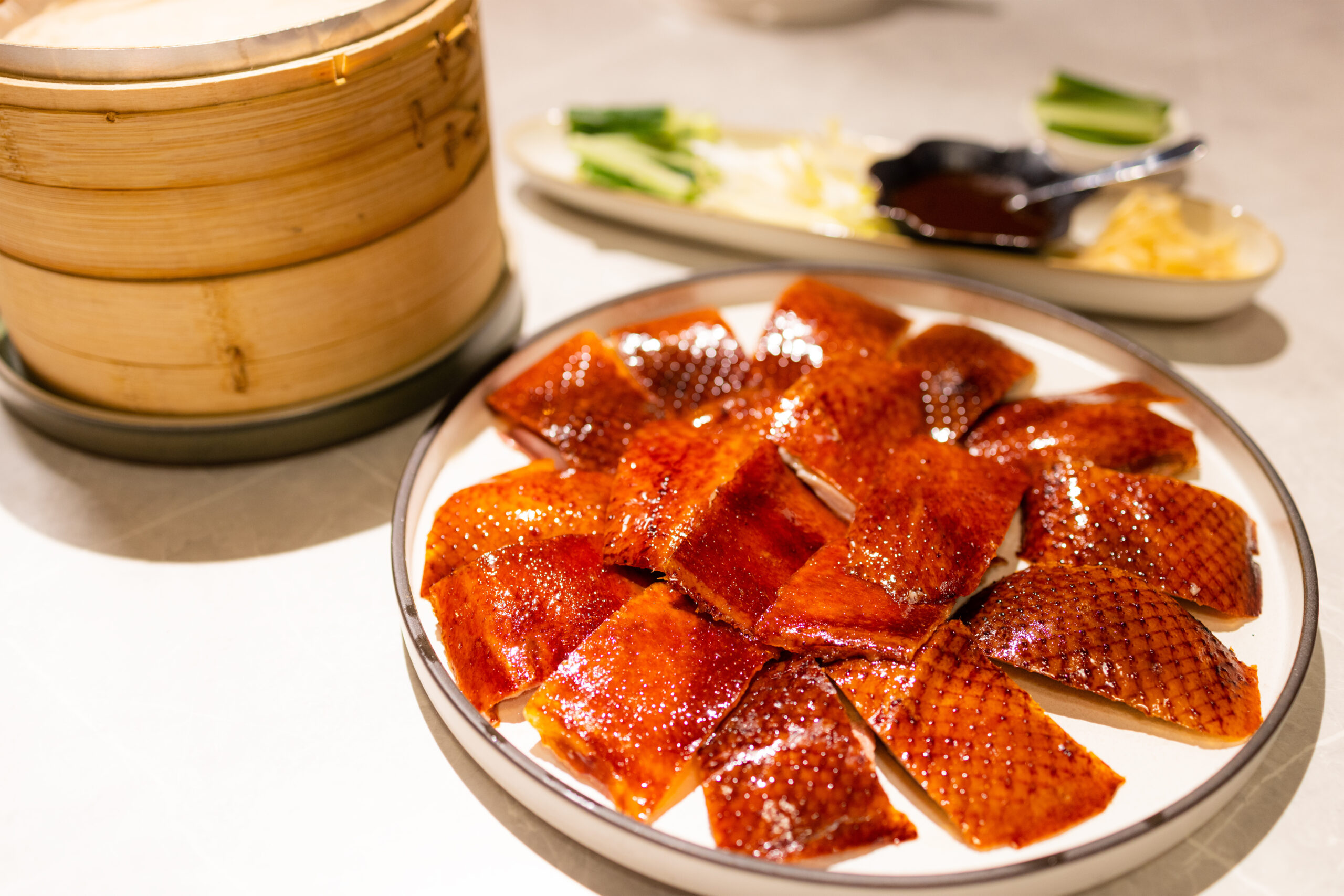 Plate of glossy roasted duck slices arranged on a white dish with a wooden steamer in the background