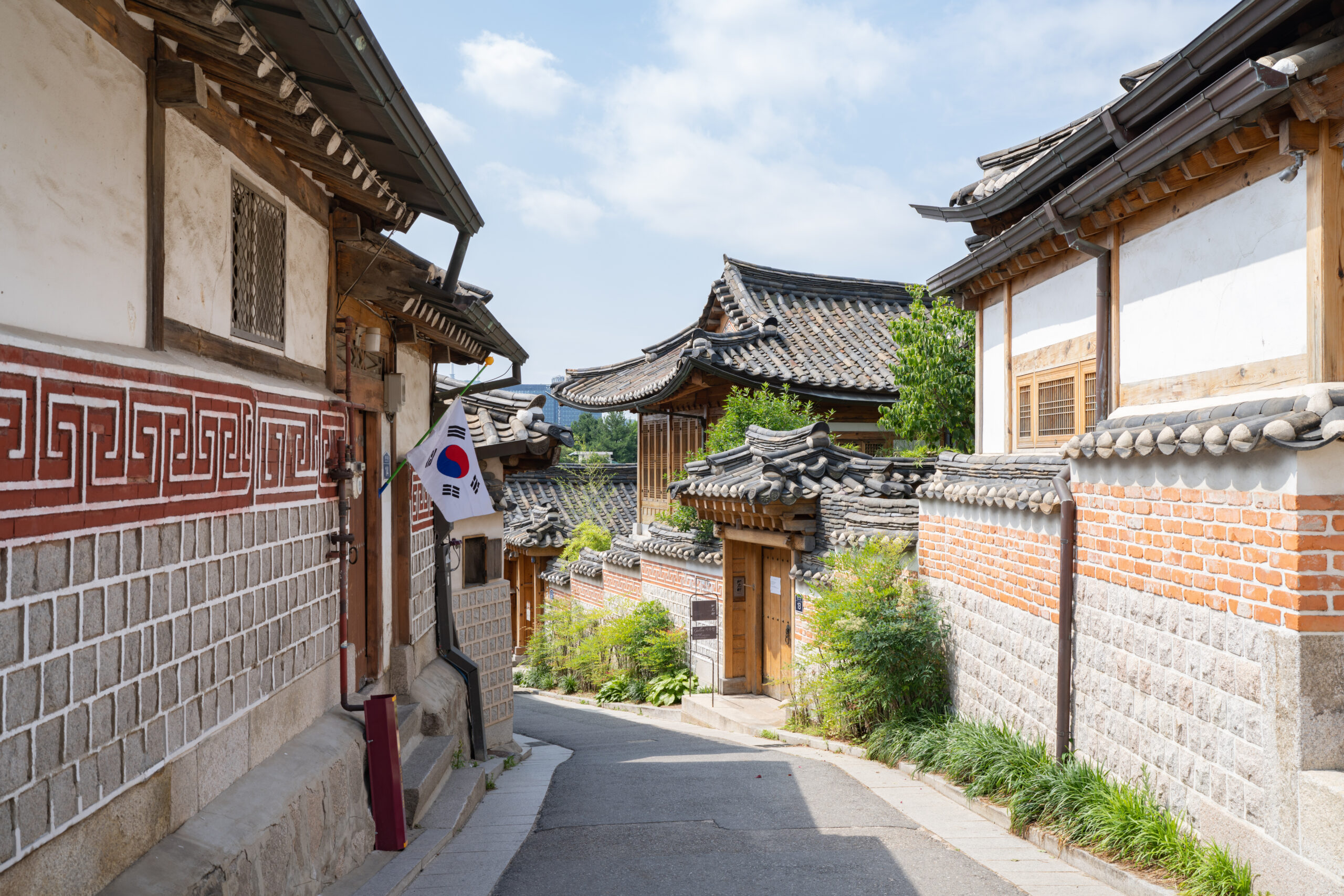 Narrow alley in a traditional Korean village with tiled roofs, white walls, and brick base walls; a South Korean flag hangs along the street.