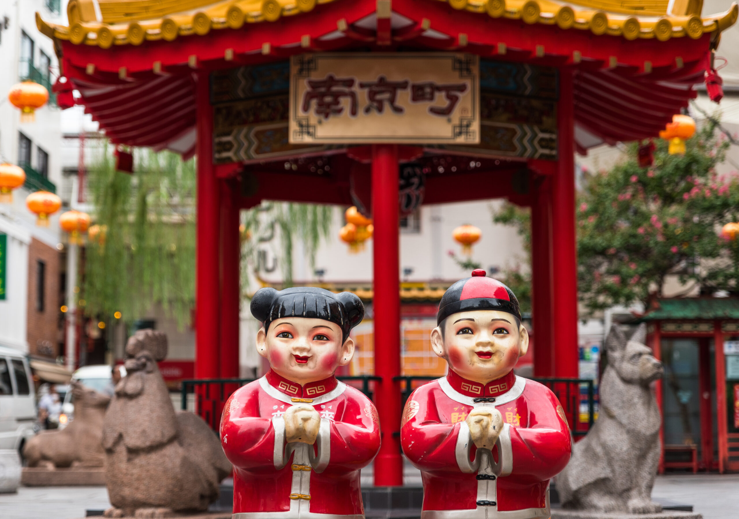 Two large red Chinese New Year statues with smiling faces stand in a street scene under a decorative red pavilion and lanterns.