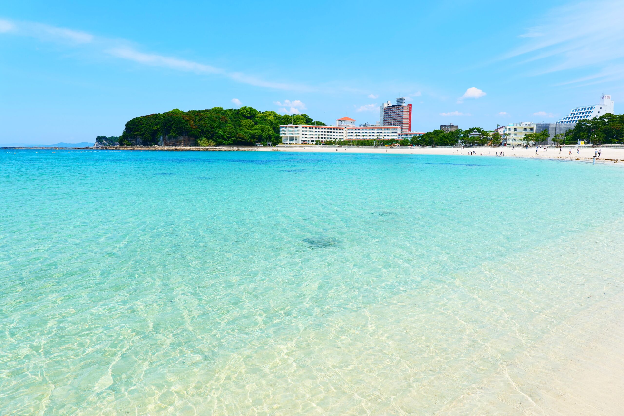 Tropical beach with clear turquoise water and buildings along the shore under a bright blue sky, horizon-wide view.