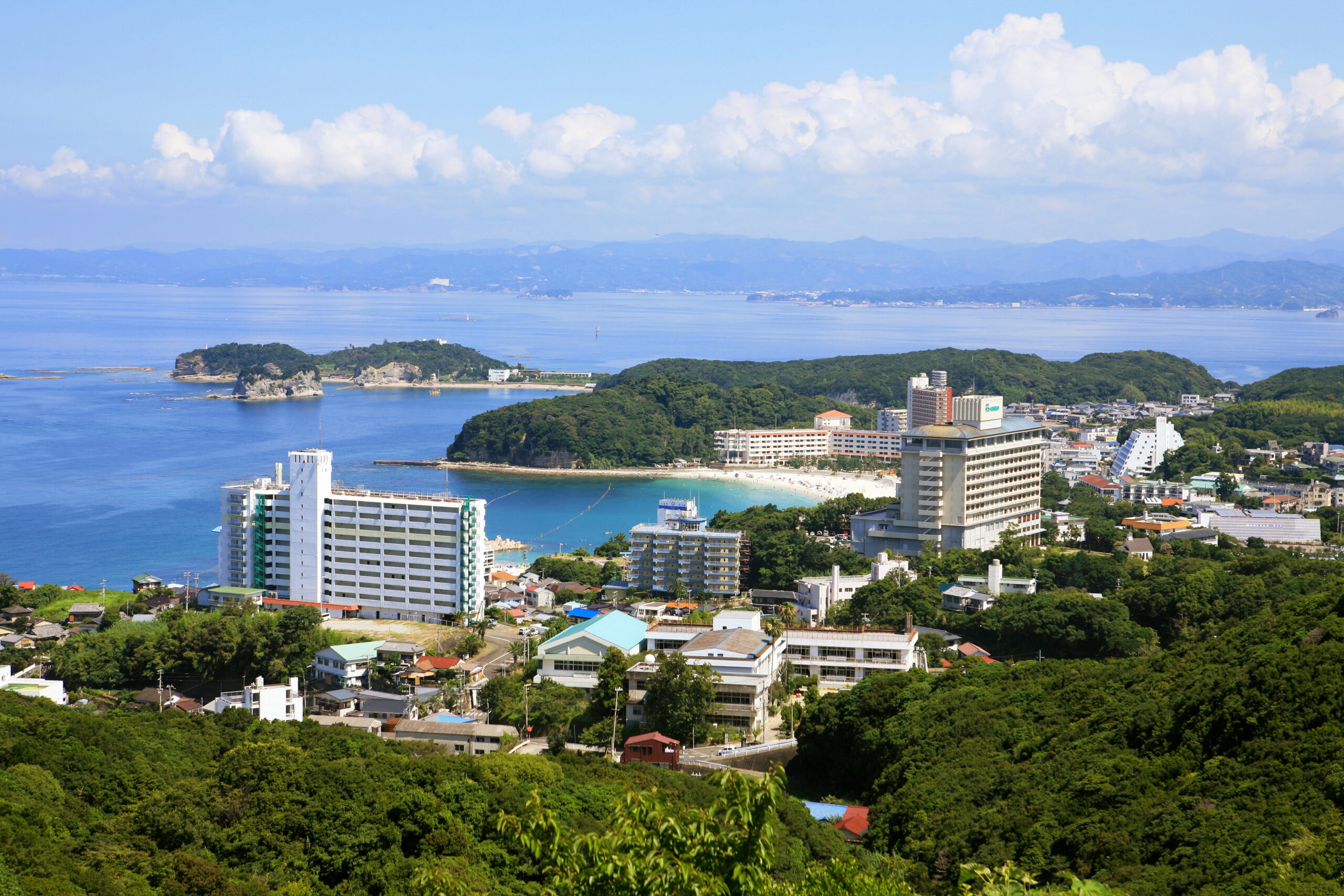 A coastal cityscape with high-rise buildings along a shoreline, green hills in the foreground, and calm blue sea extending to distant mountains under a cloudy sky.