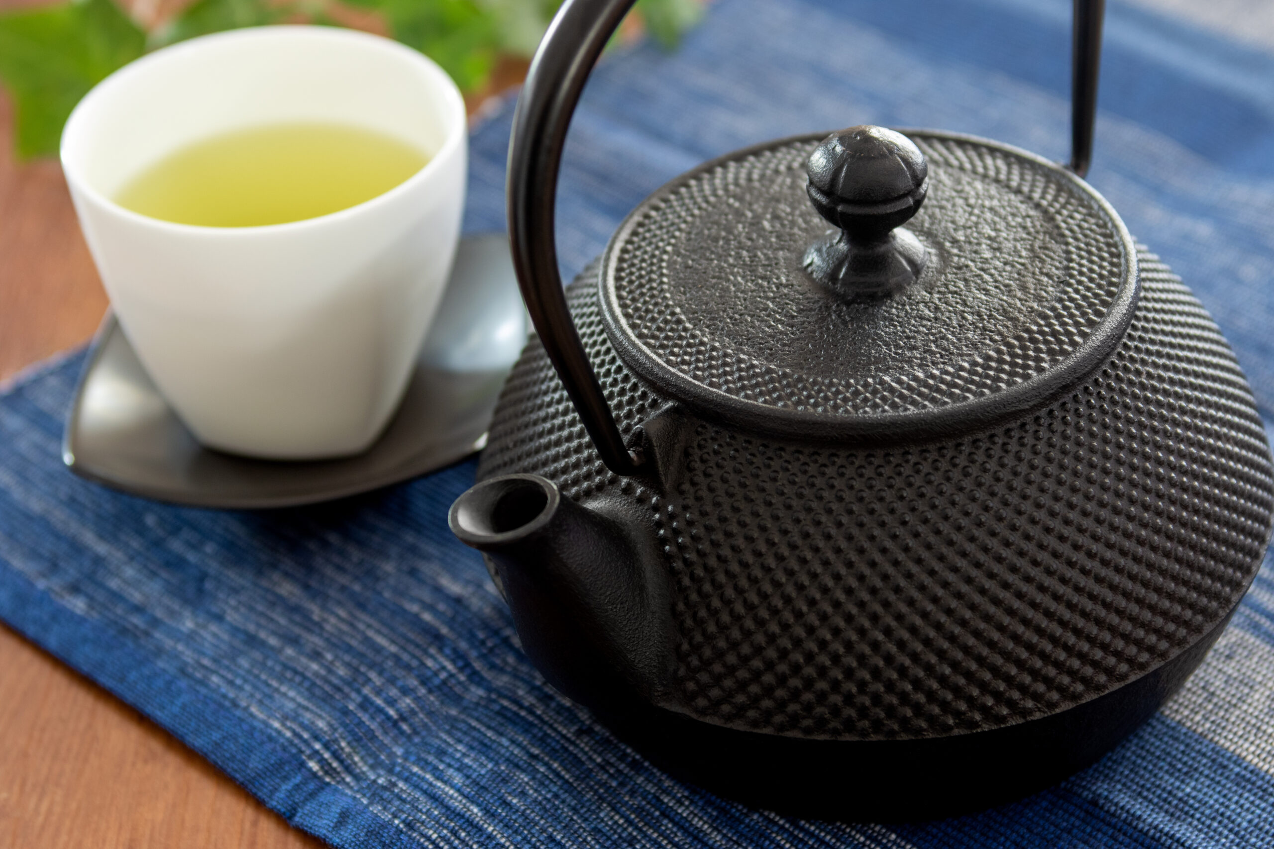 Cast iron teapot with a textured surface beside a white cup of pale yellow tea on a blue fabric placemat/tablecloth.