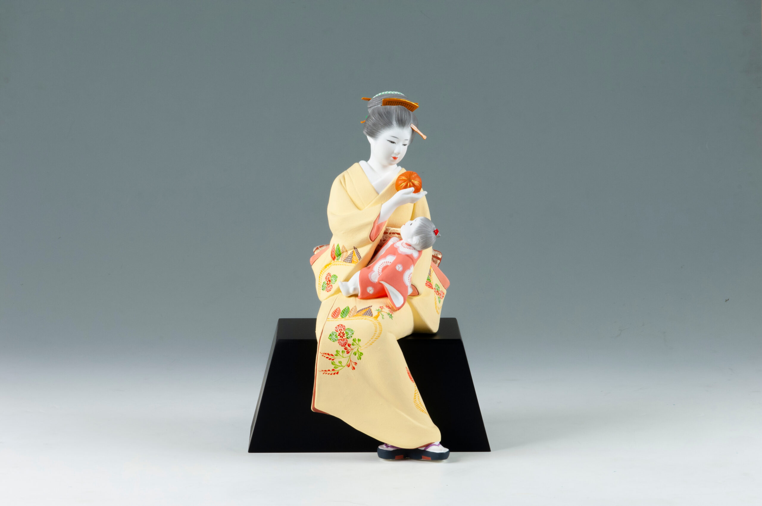 Traditional Japanese geisha doll in a pale yellow kimono with floral embroidery, seated on a black display base and holding an orange ball-like object.
