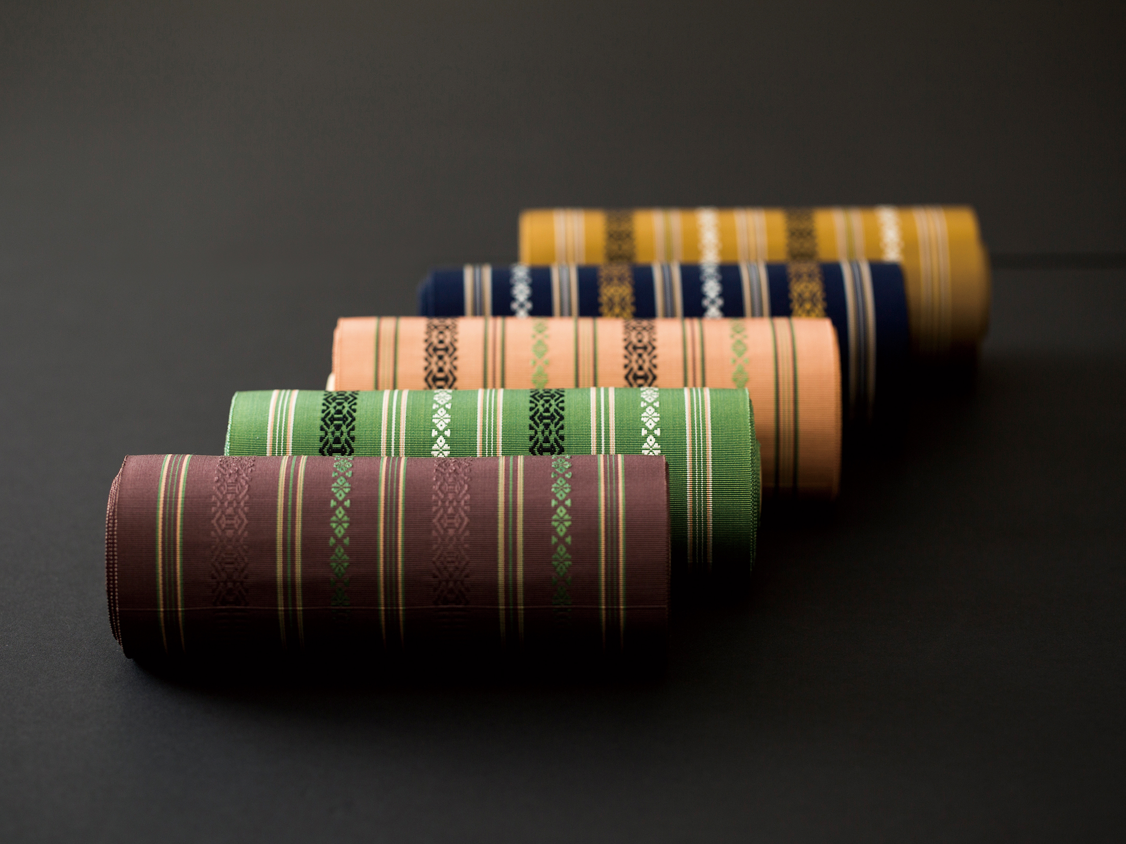 Stacked rolled woven textiles in green, pink, blue, yellow, and purple with decorative white and black patterns on a dark background.