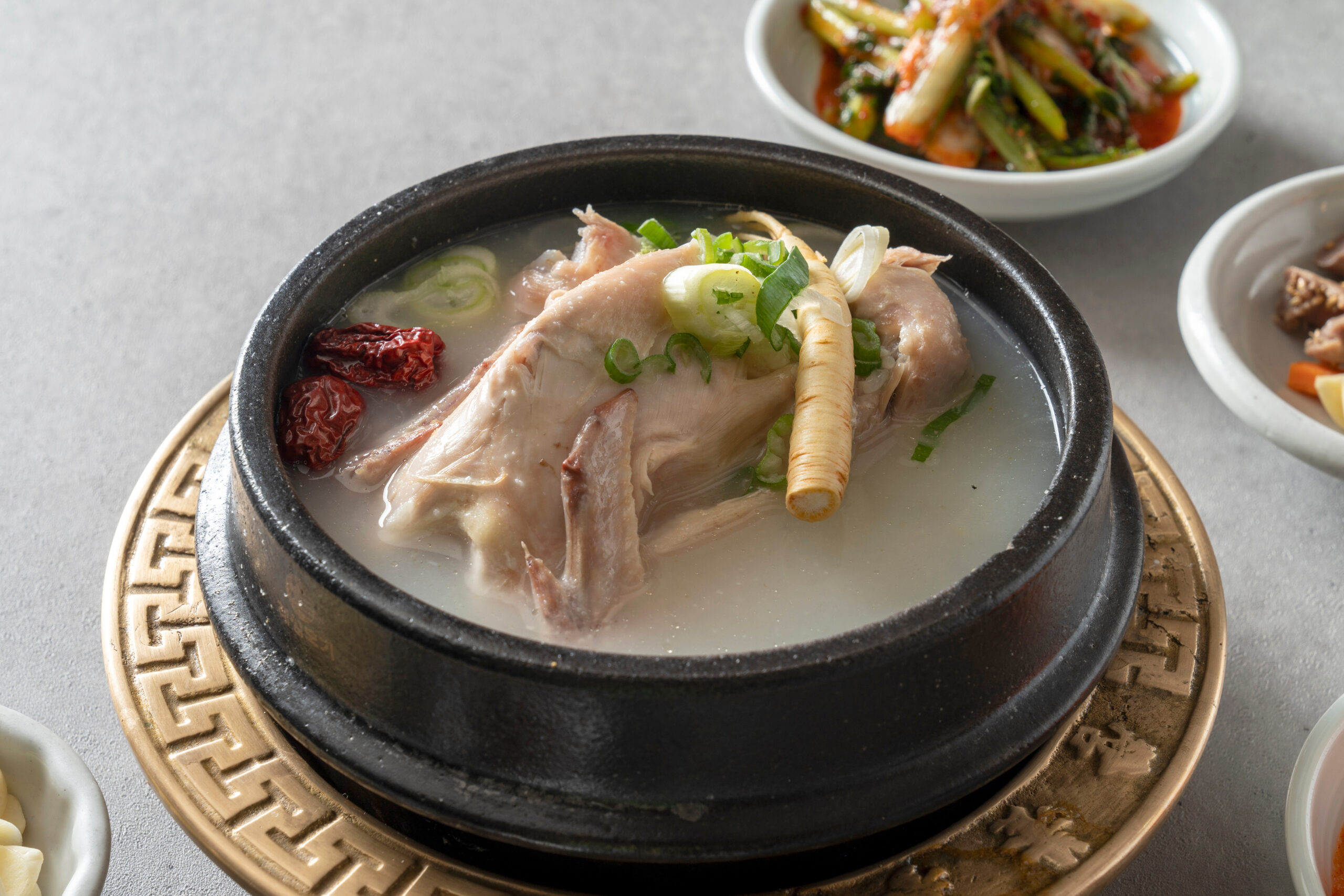 Hot Korean ginseng chicken soup in a black ceramic pot, topped with green onions and a ginseng root, with side dishes nearby.