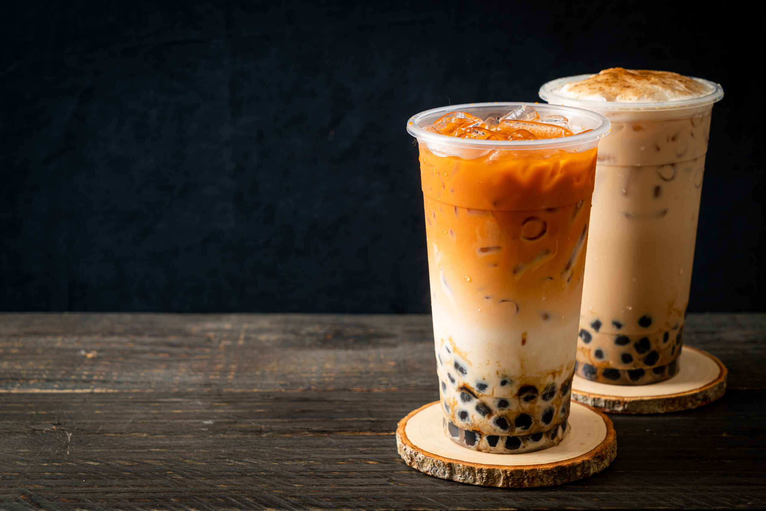 Two iced tea beverages with tapioca pearls (boba) stacked on wooden coasters against a dark background, one with orange color and ice.