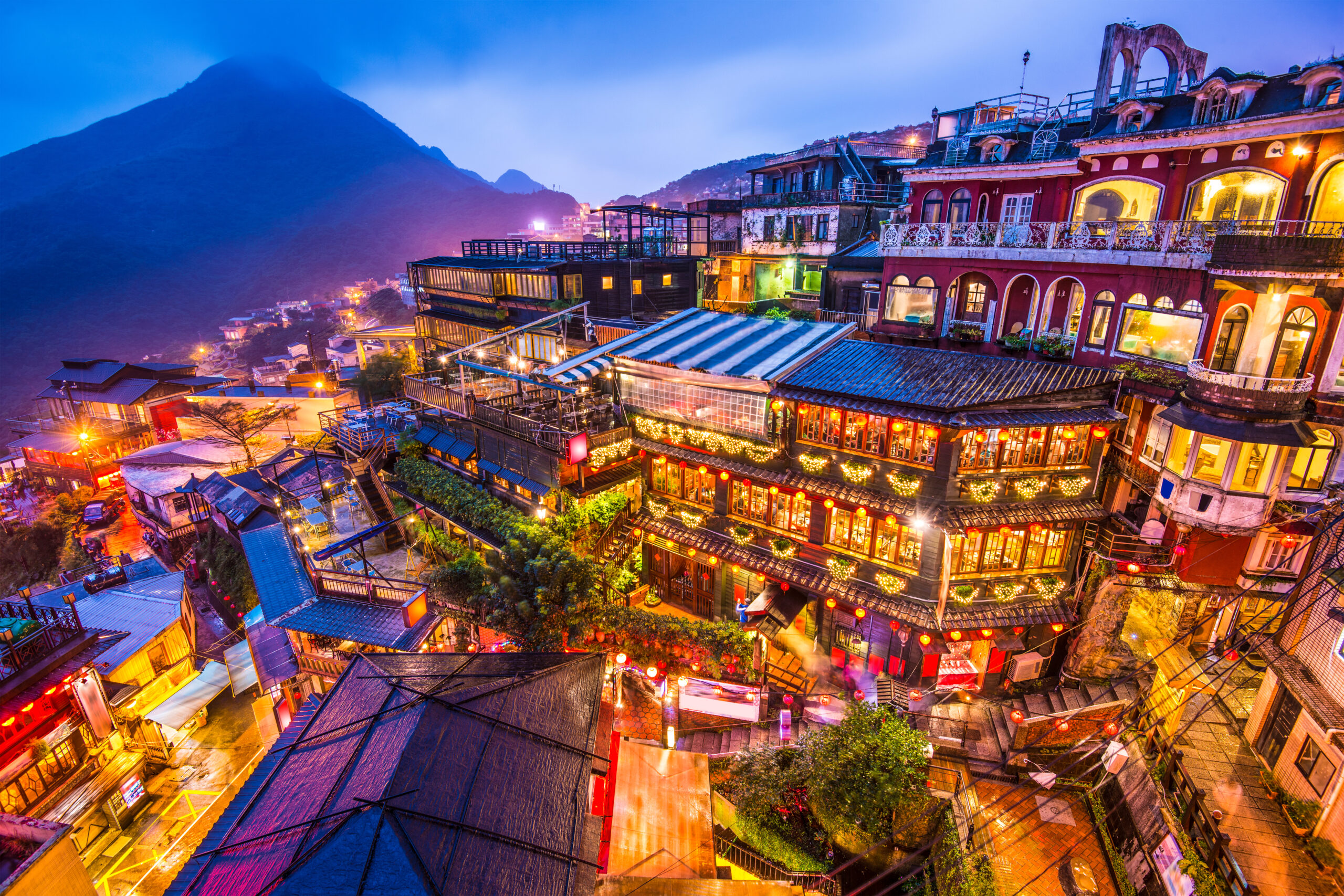 Nighttime aerial view of a colorful hillside town with lit balconies, lanterns, and glowing windows against a blue mountain backdrop