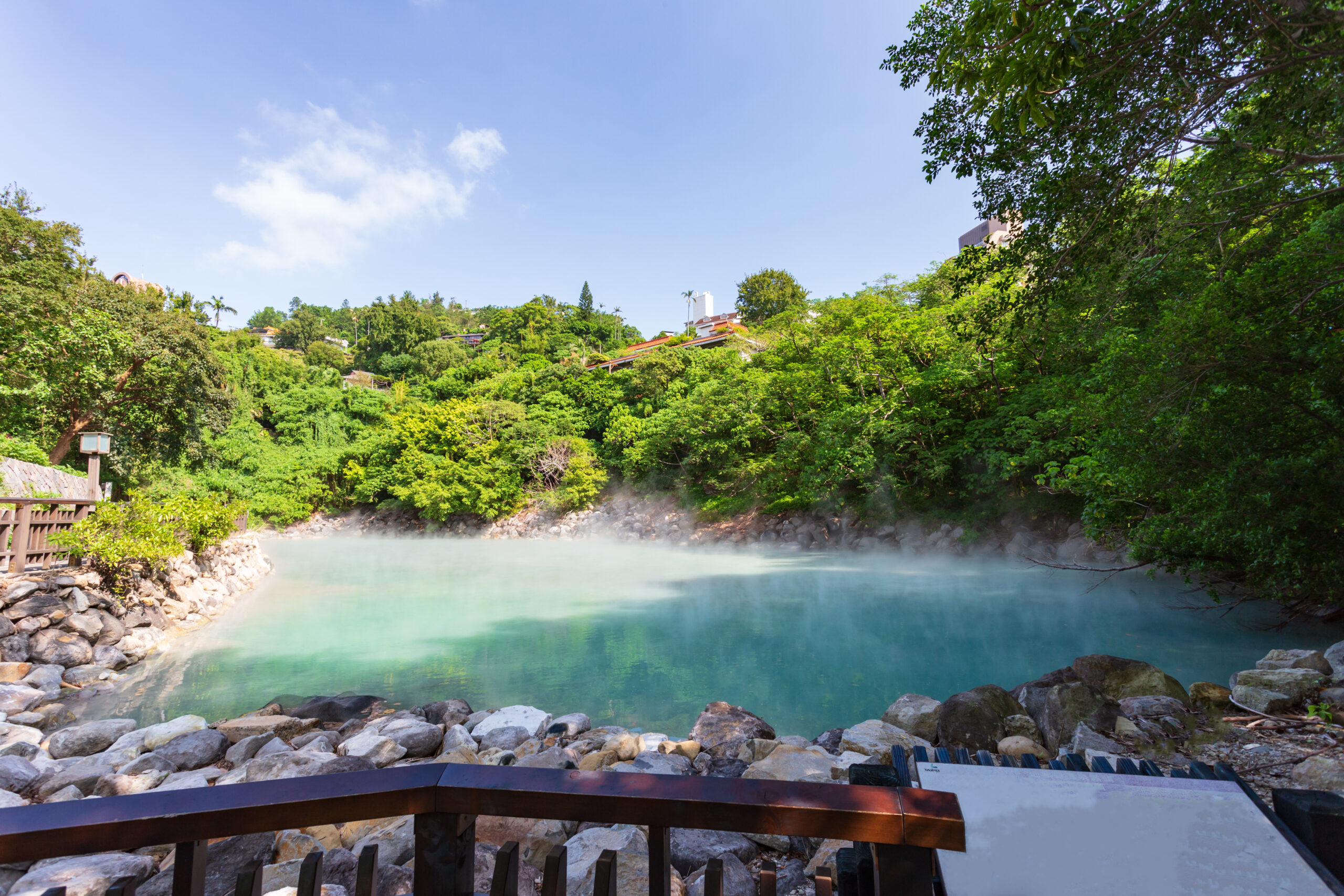 Wooden railing in the foreground overlooks a rocky riverbank, turquoise water, and a lush green hillside with houses under a bright blue sky.