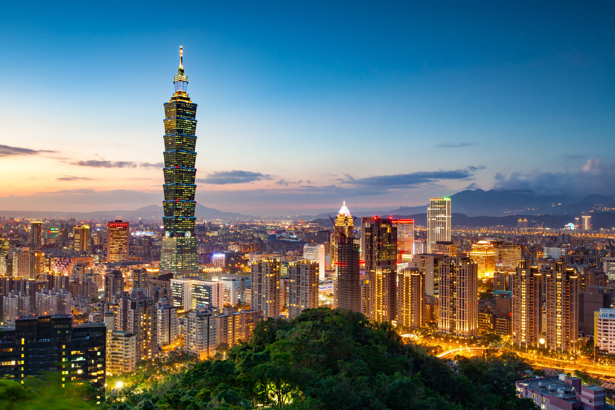 Evening cityscape featuring Taipei 101 towering over illuminated high-rise buildings and distant mountains in the background.