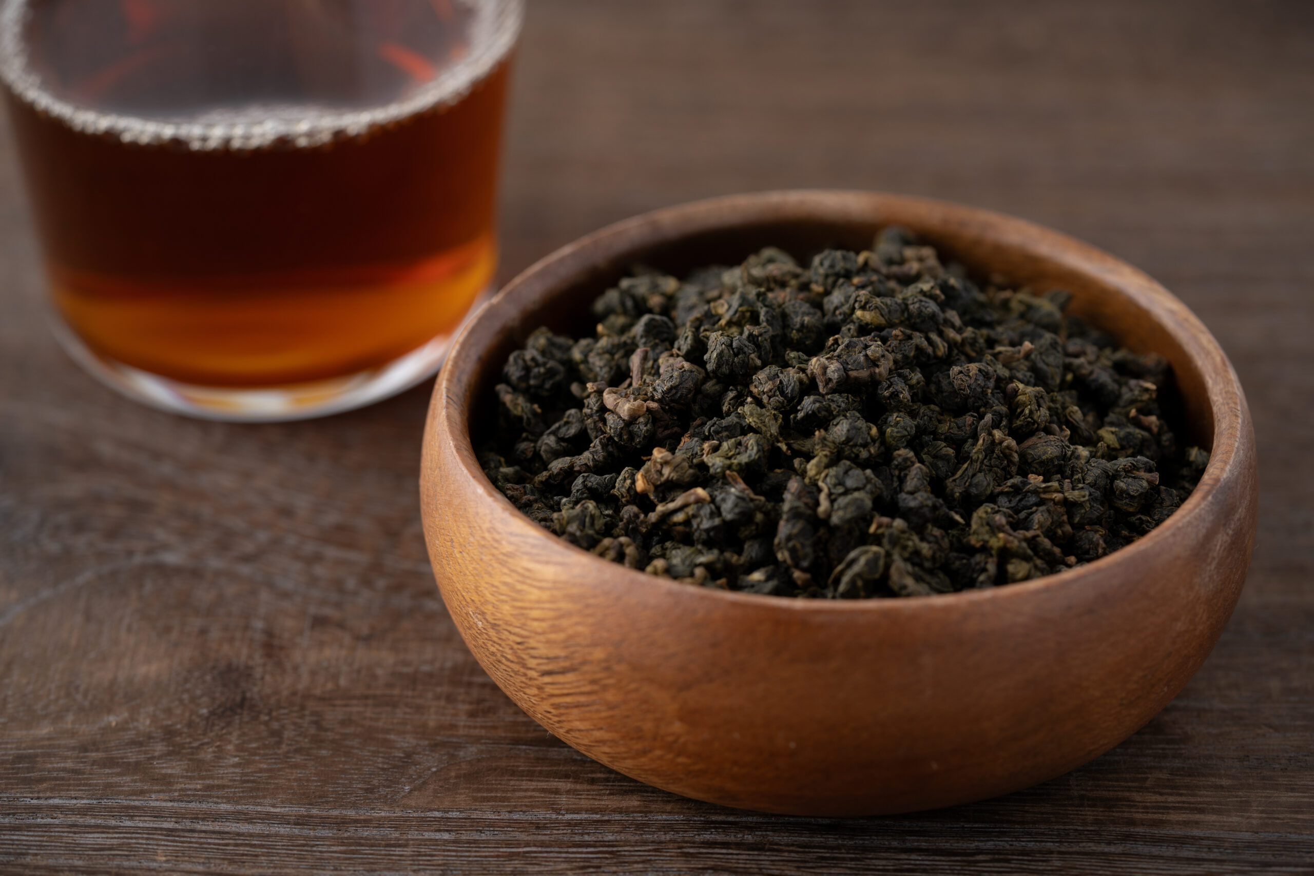 Wooden bowl filled with loose tea leaves on a rustic wooden table, with a glass of brewed tea in the blurred background.
