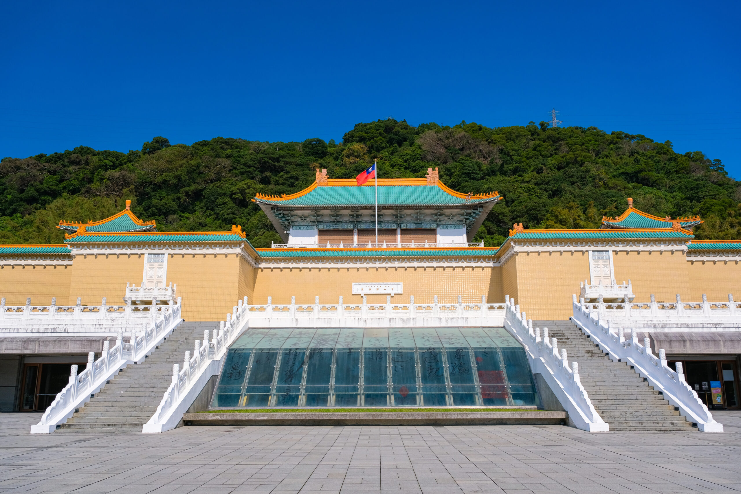 Temple entrance with green tiled rooftops and orange trim, white stone stairs, and a flagpole in front against a forest hill.