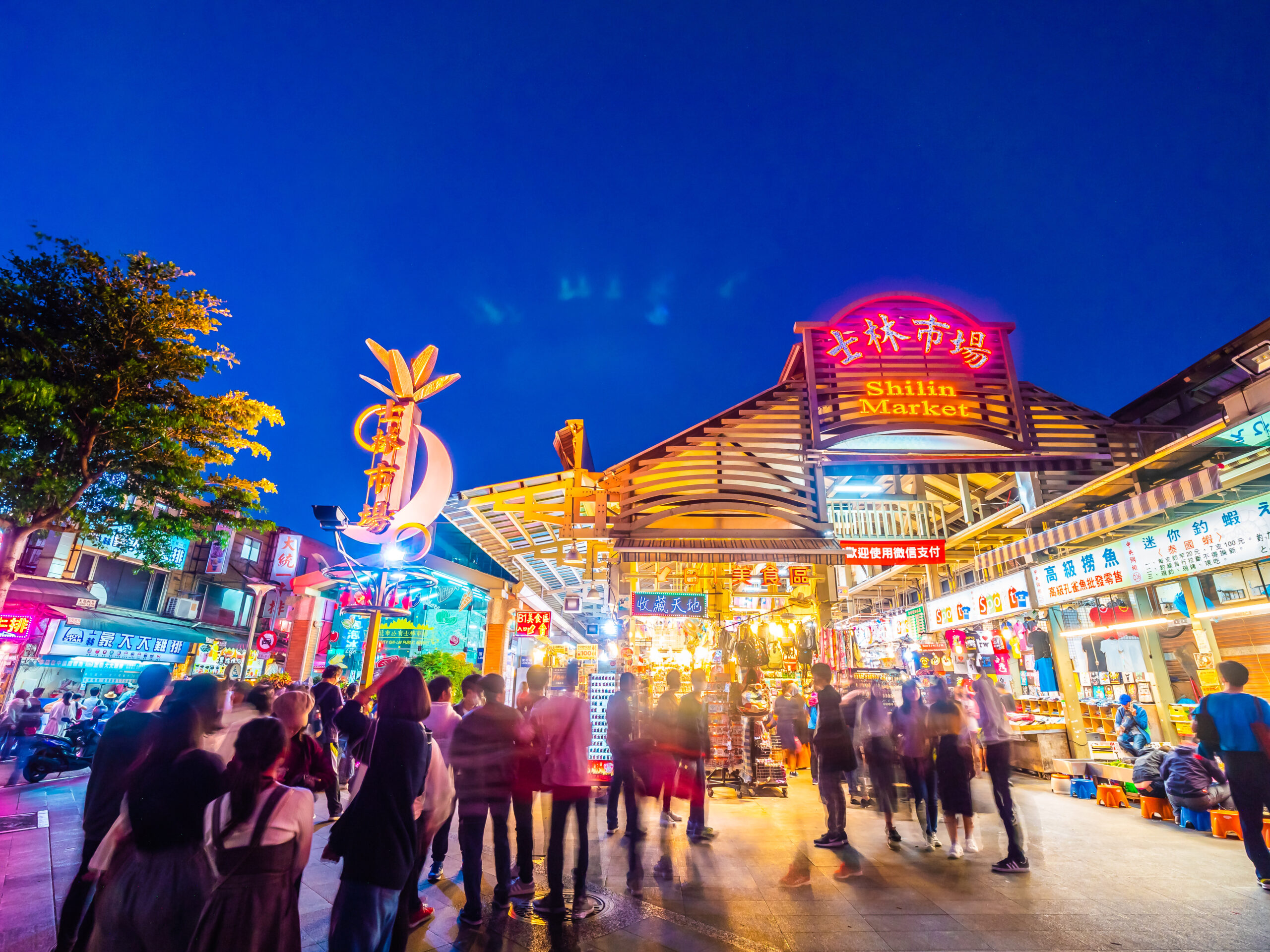 Nighttime view of Shilin Market entrance with neon sign, colorful stalls, and a crowded walkway full of shoppers.