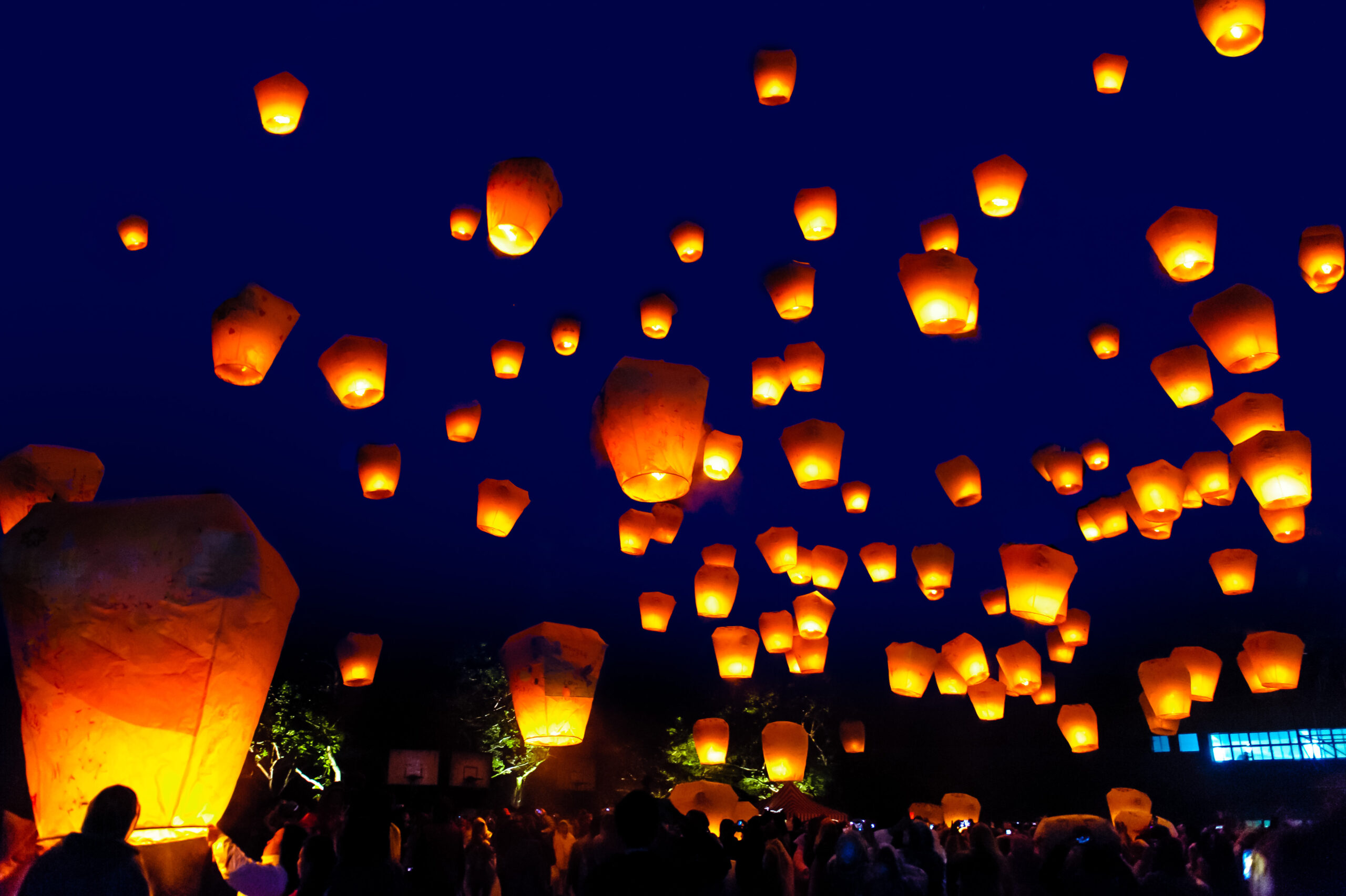 Night sky filled with glowing orange lanterns floating upward during a festival, with a crowd visible below at night.