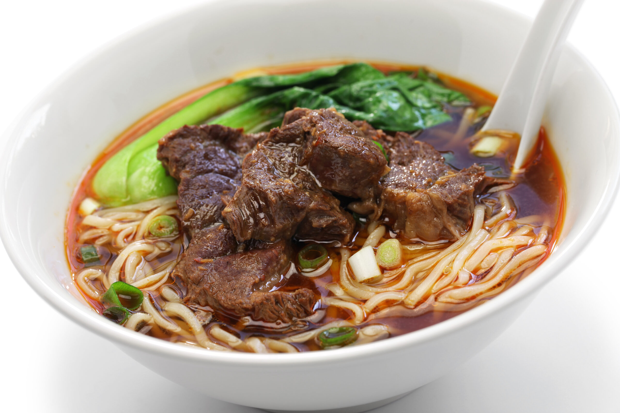Bowl of beef noodle soup with leafy greens and scallions in a reddish broth.