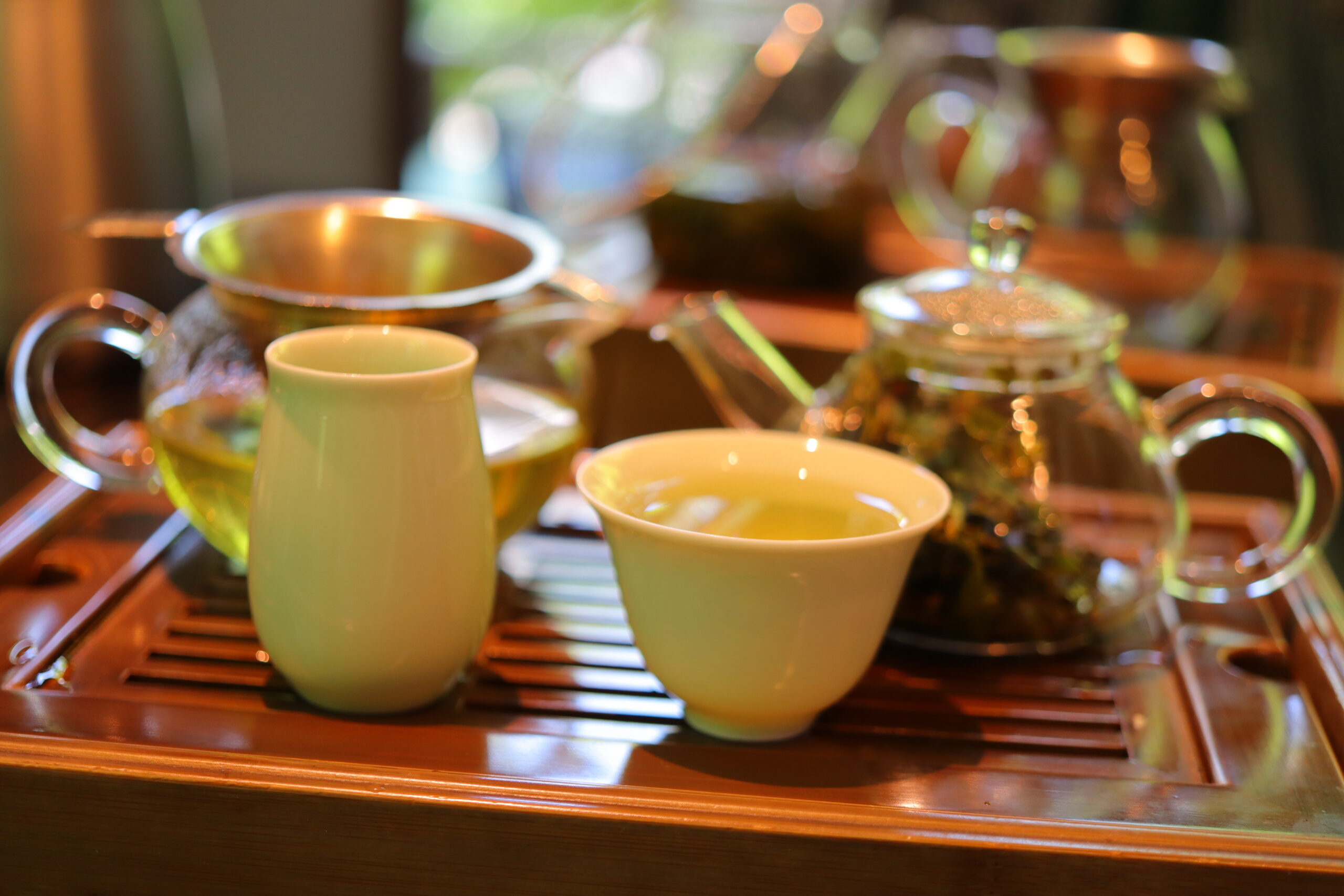 Green ceramic cups and a glass teapot sit on a wooden tray, with a warmly blurred kitchen background.
