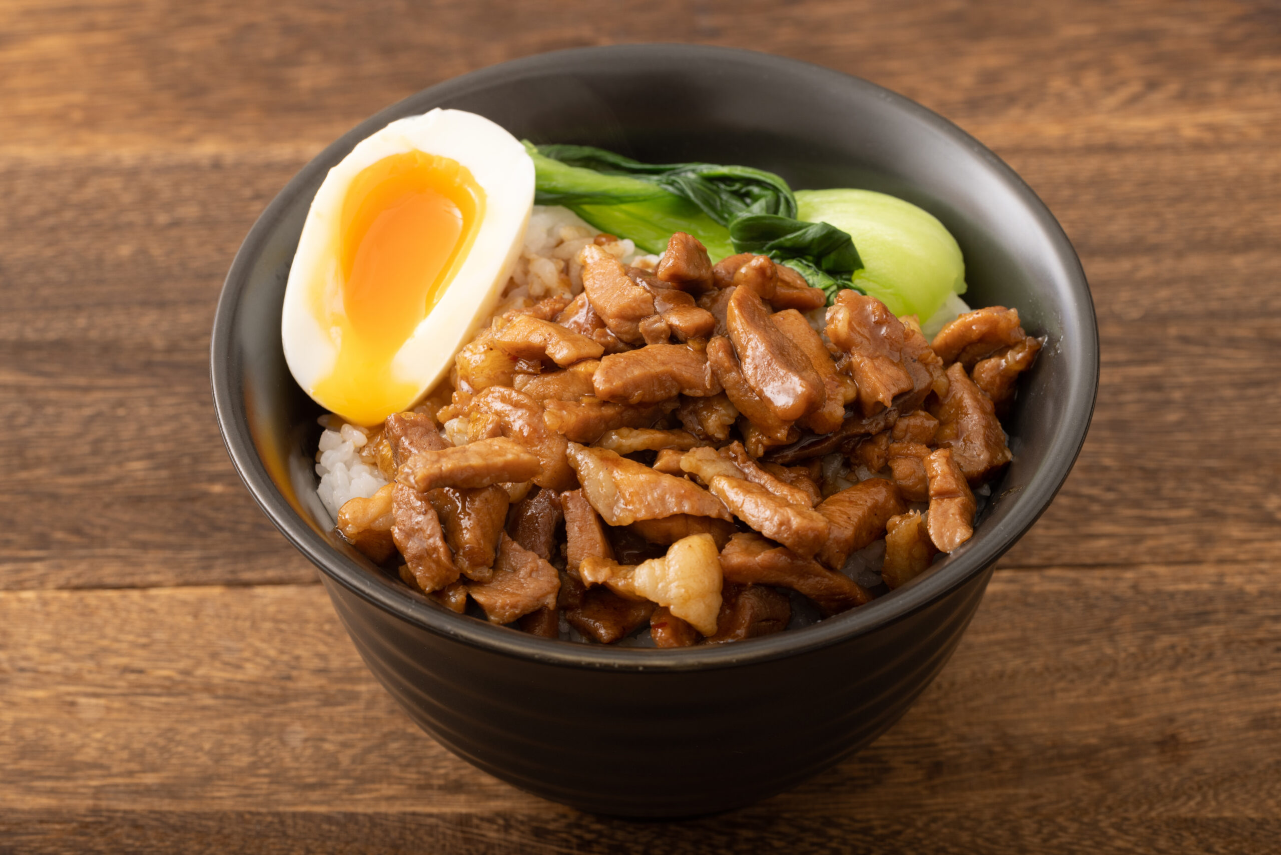 Bowl of braised pork over rice with a halved soft-cooked egg and bok choy on top, in a black bowl.