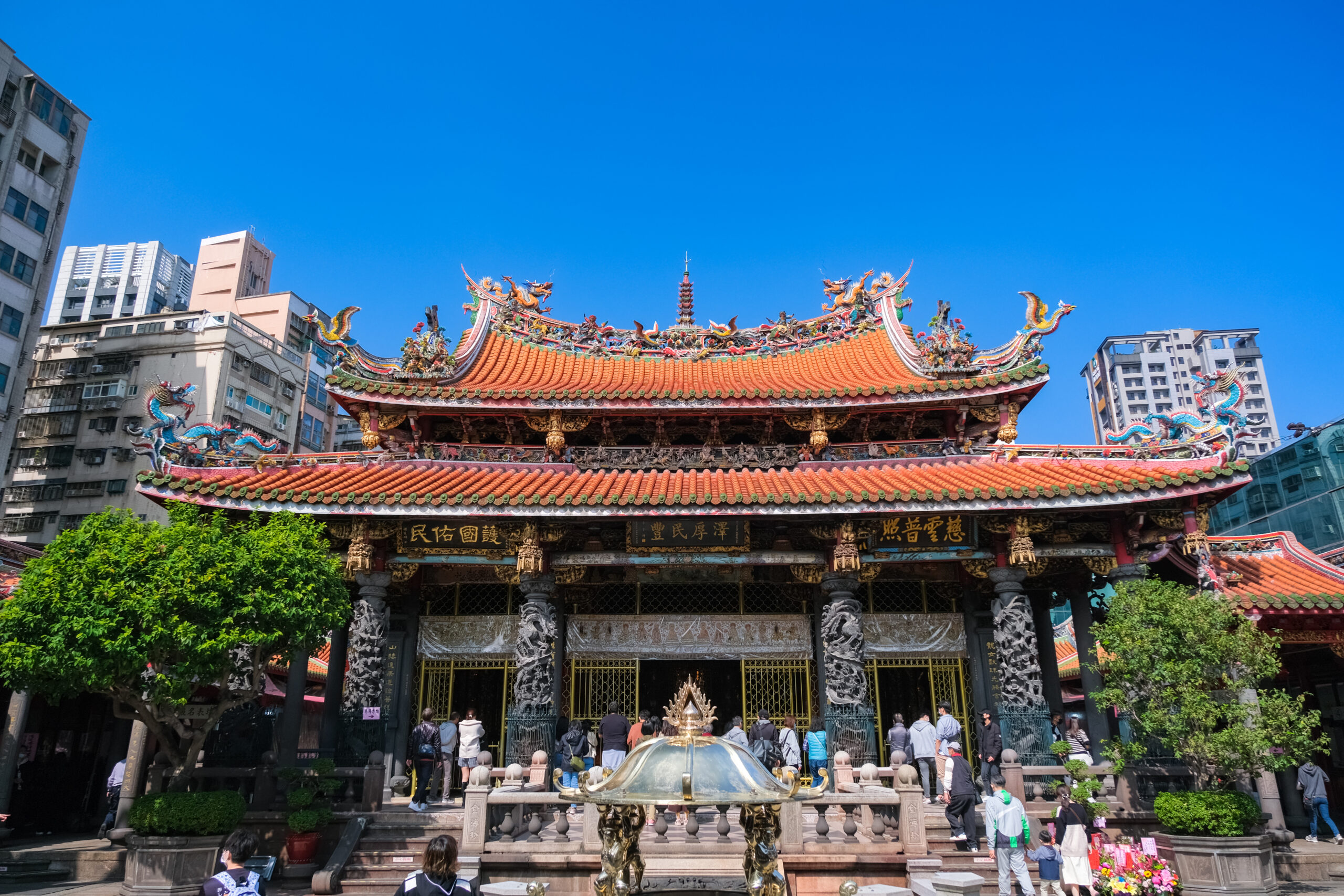 Ornate Asian temple with a red tiled roof and dragon sculptures against a clear blue sky, surrounded by modern buildings.