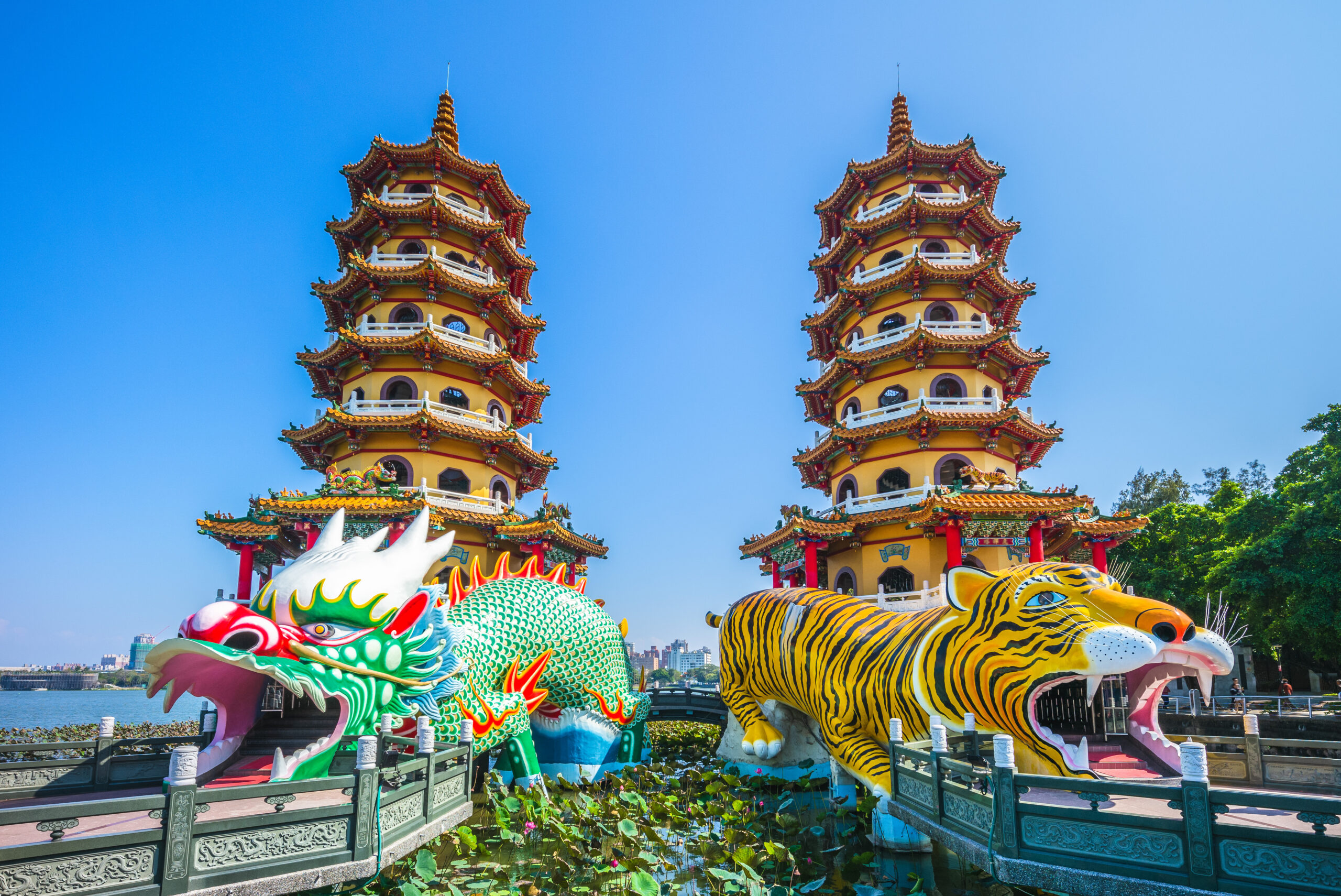 Two ornate Chinese pagodas rise side by side against a clear blue sky, flanked by colorful dragon and tiger floats at a water-side temple complex.