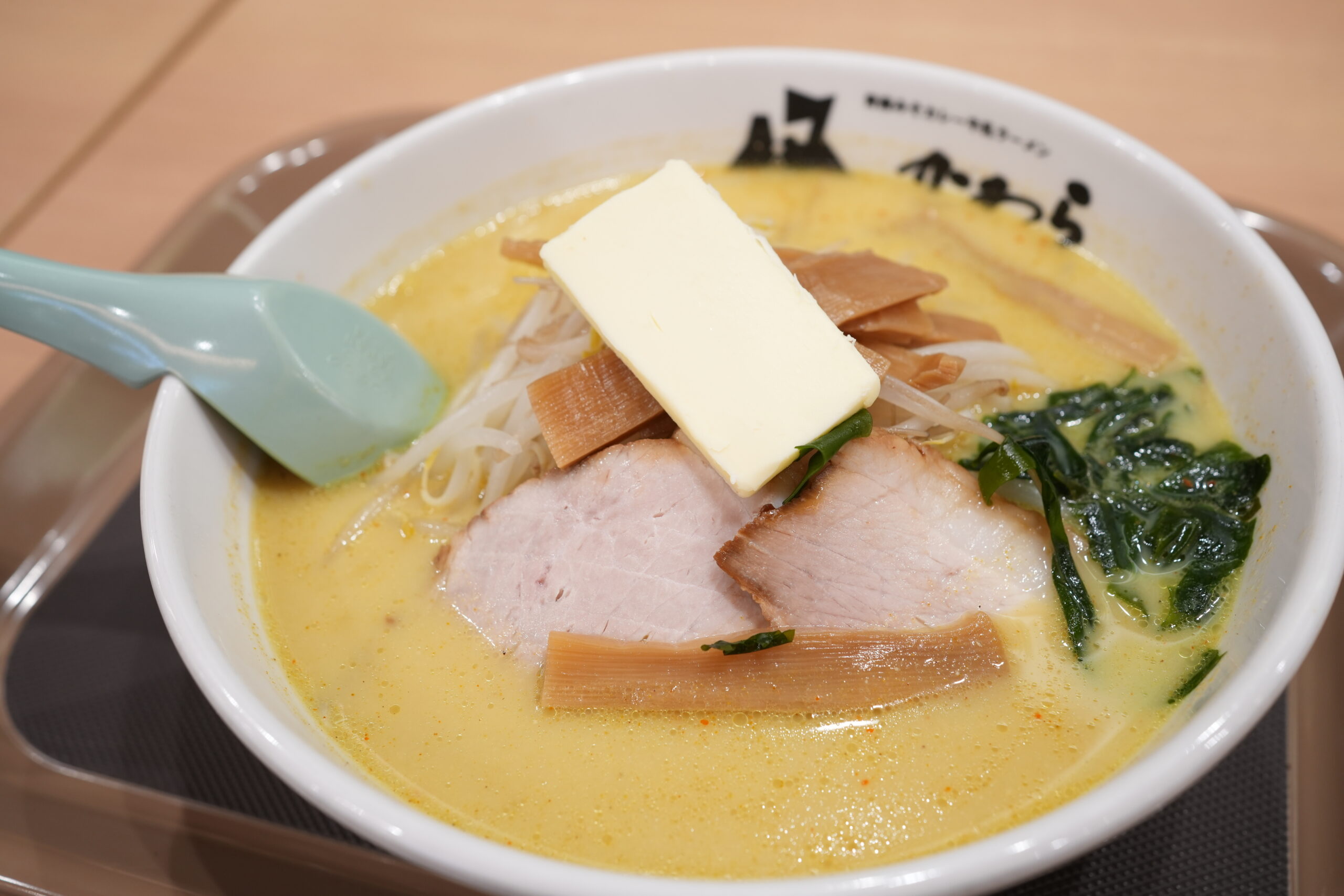 Creamy yellow ramen broth in a white bowl topped with a pat of butter, slices of pork, bamboo shoots, and greens with noodles visible underneath.
