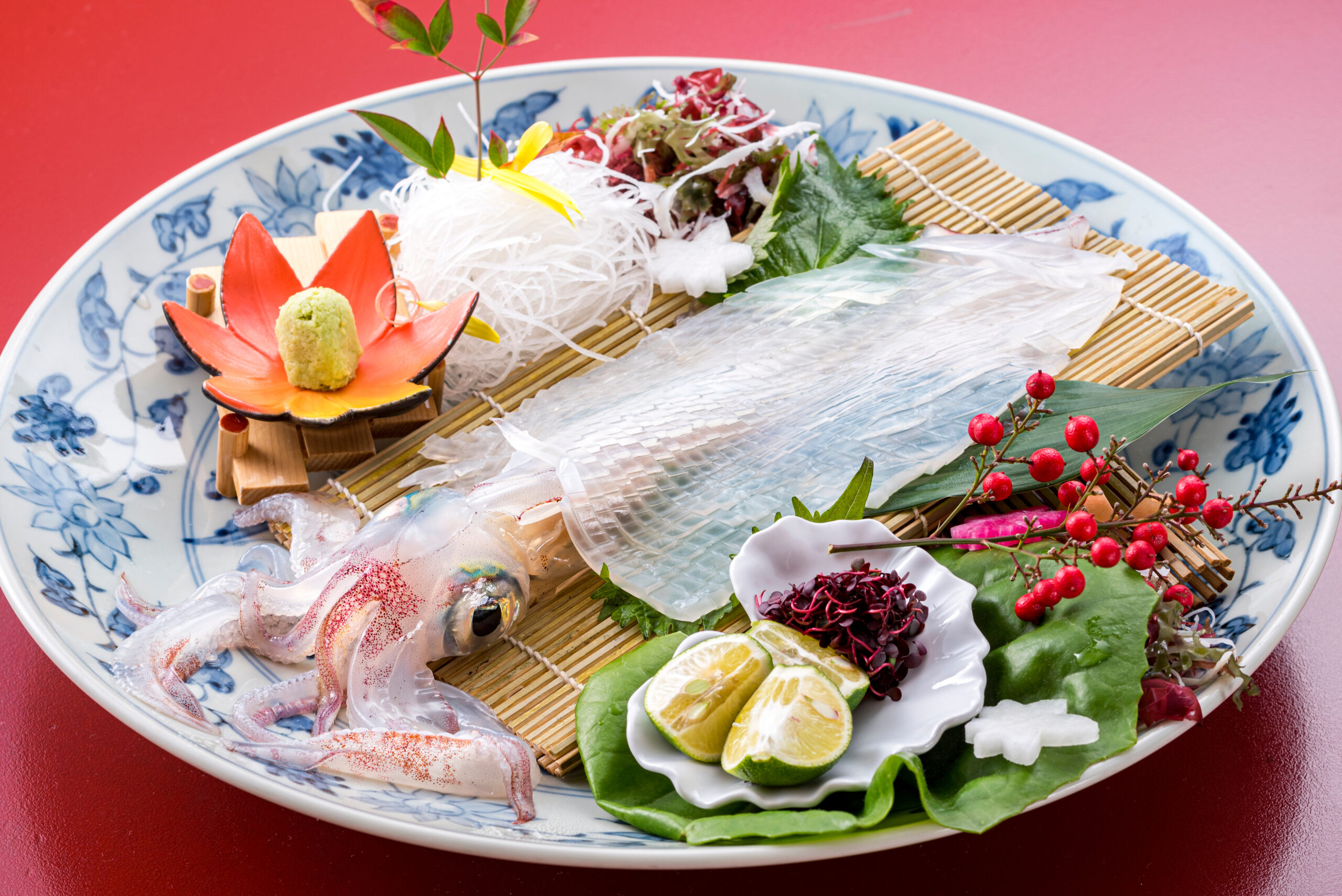 Assorted sashimi on a blue floral plate with lemon wedges, greens, and red berries against a red background.
