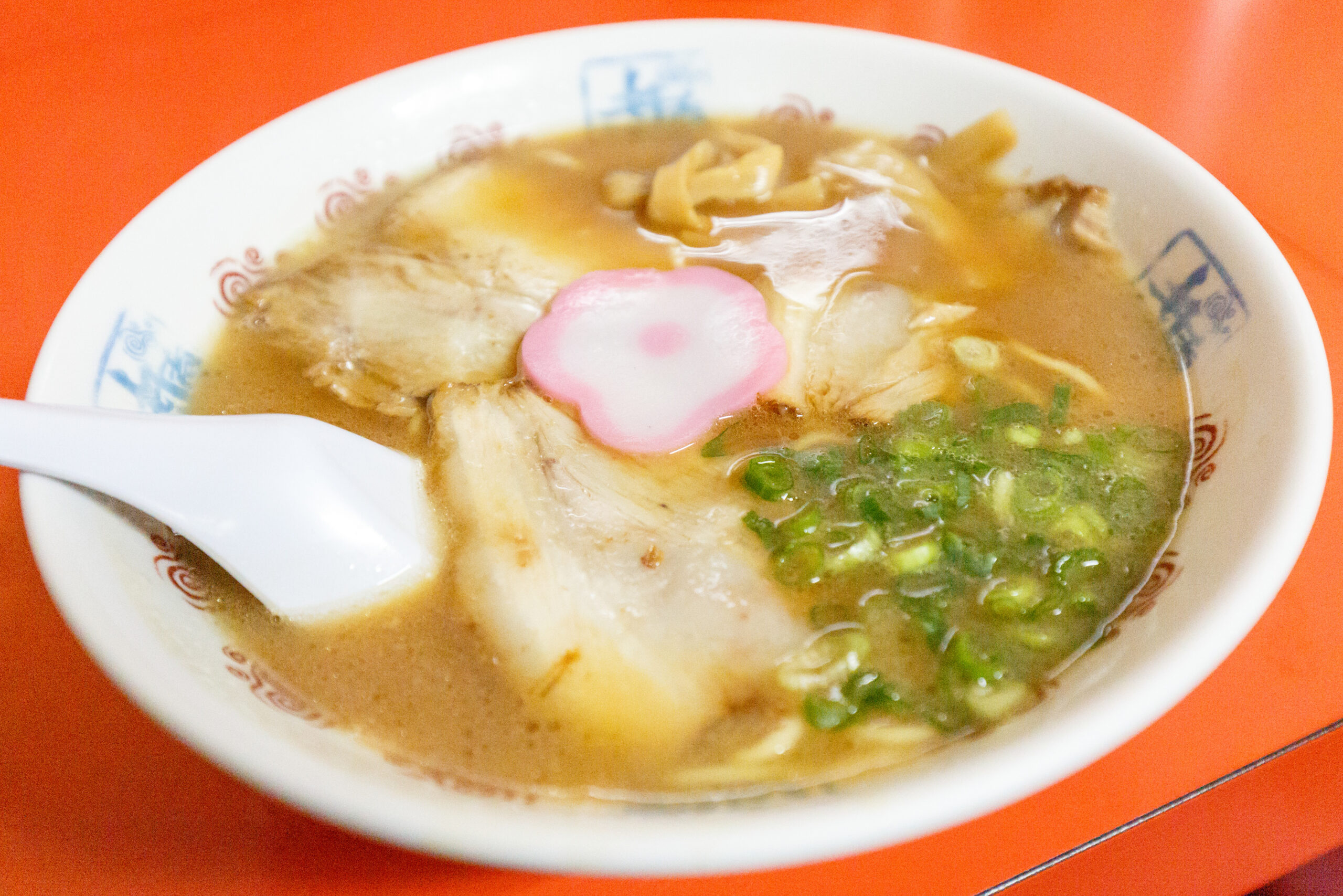 Bowl of ramen with sliced pork, pink-and-white narutomaki, green onions, and broth on an orange table.
