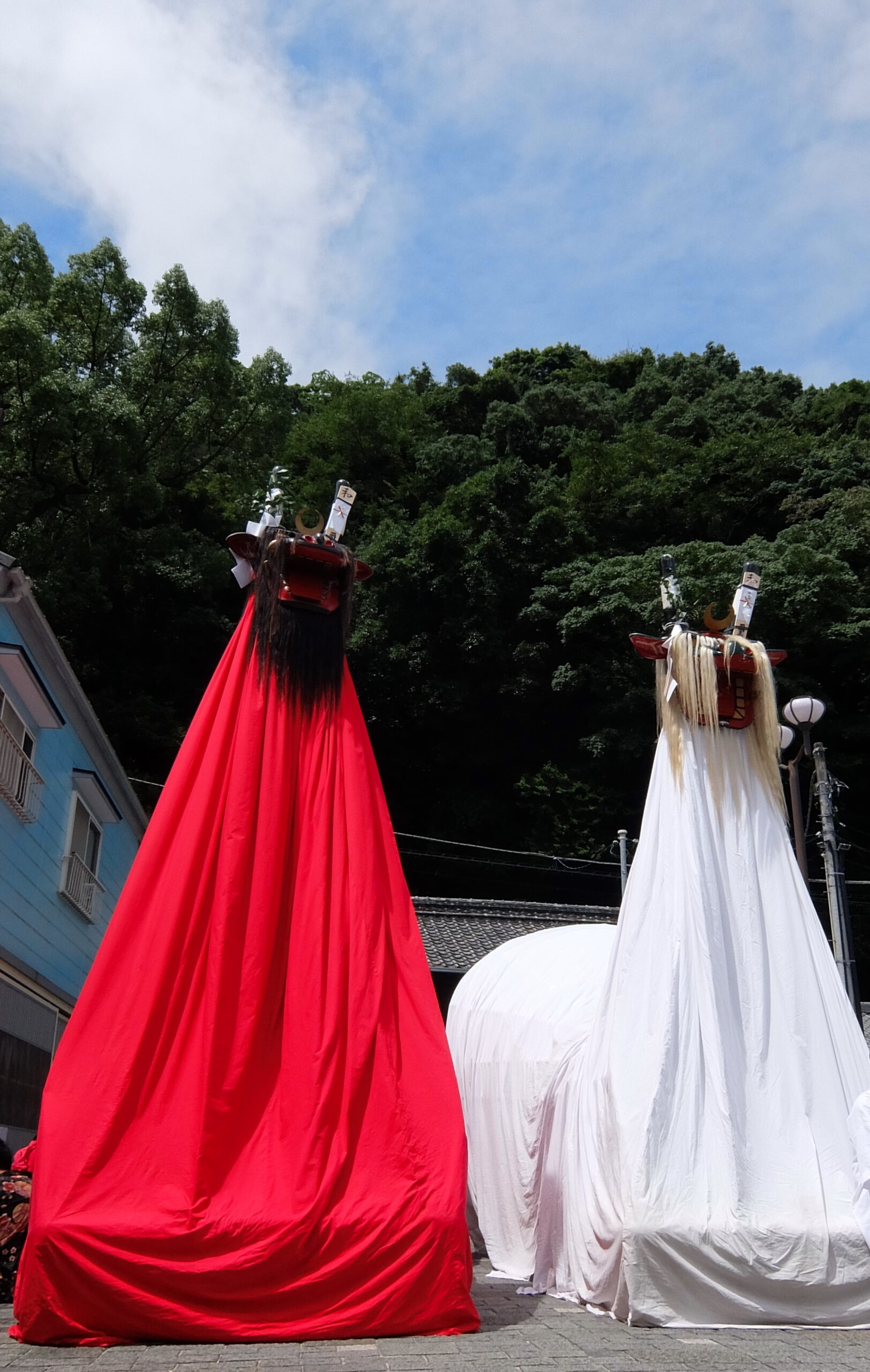 Two towering festival figures draped in bright red and white fabric with carved wooden heads, standing on a street under a blue sky.