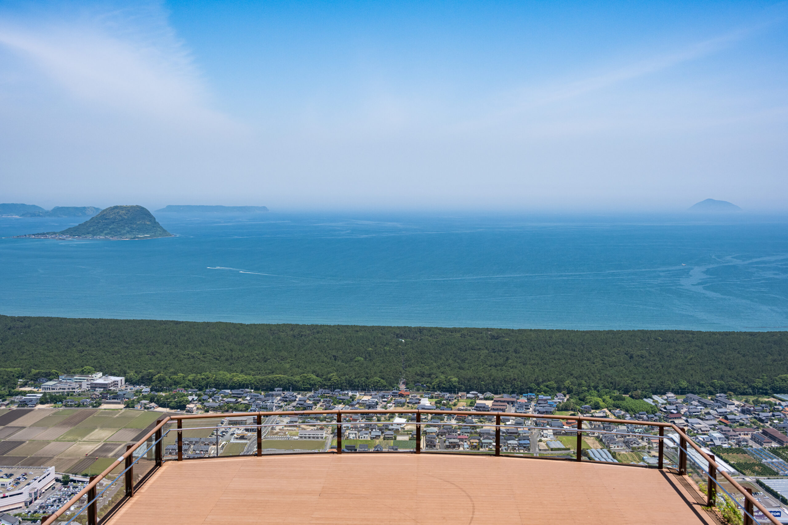 Terrace railing in the foreground overlooking a coastal town, dense forest, and a calm blue sea with distant islands on the horizon.