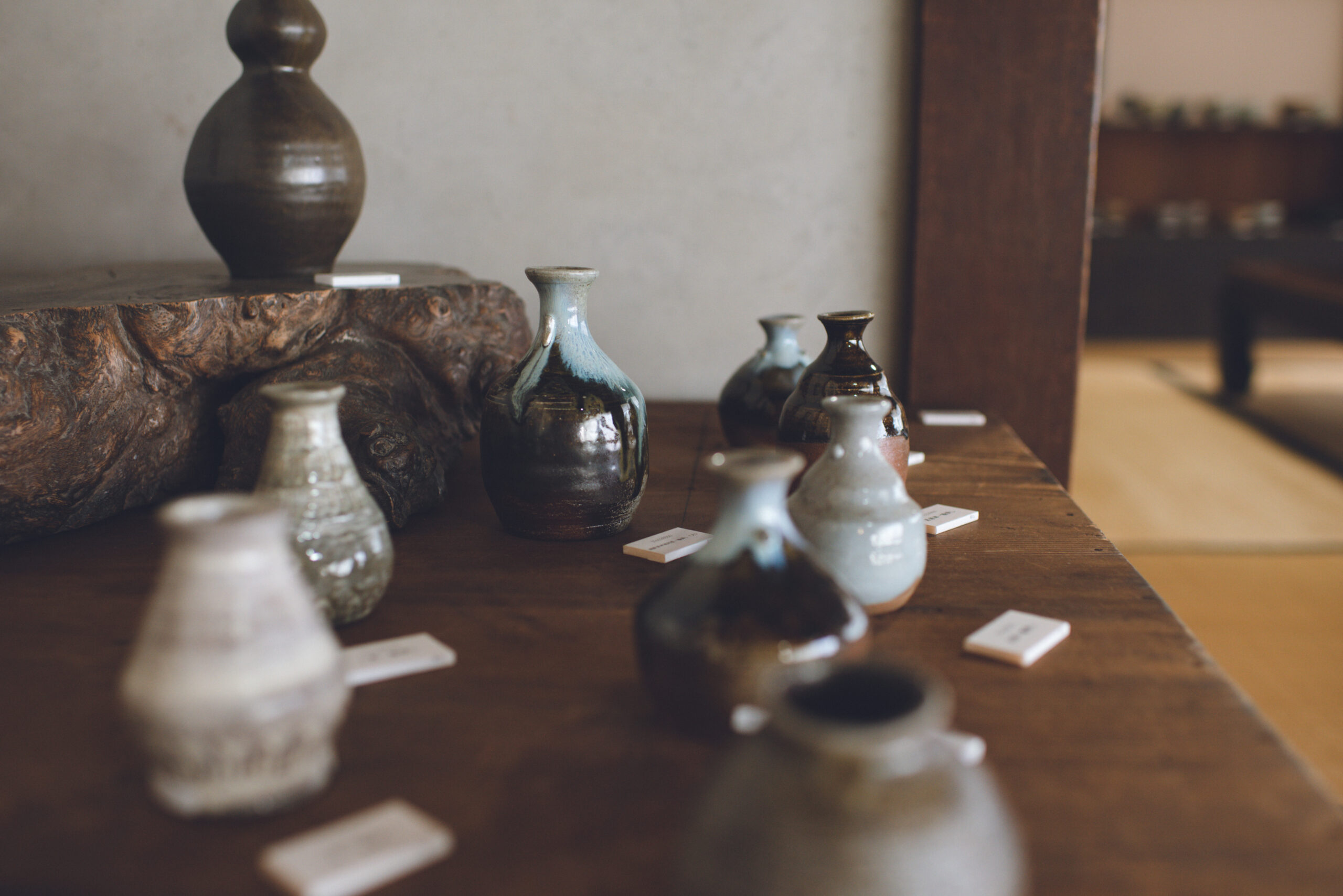 Row of ceramic vases and bottles on a wooden table in a softly lit gallery setting.
