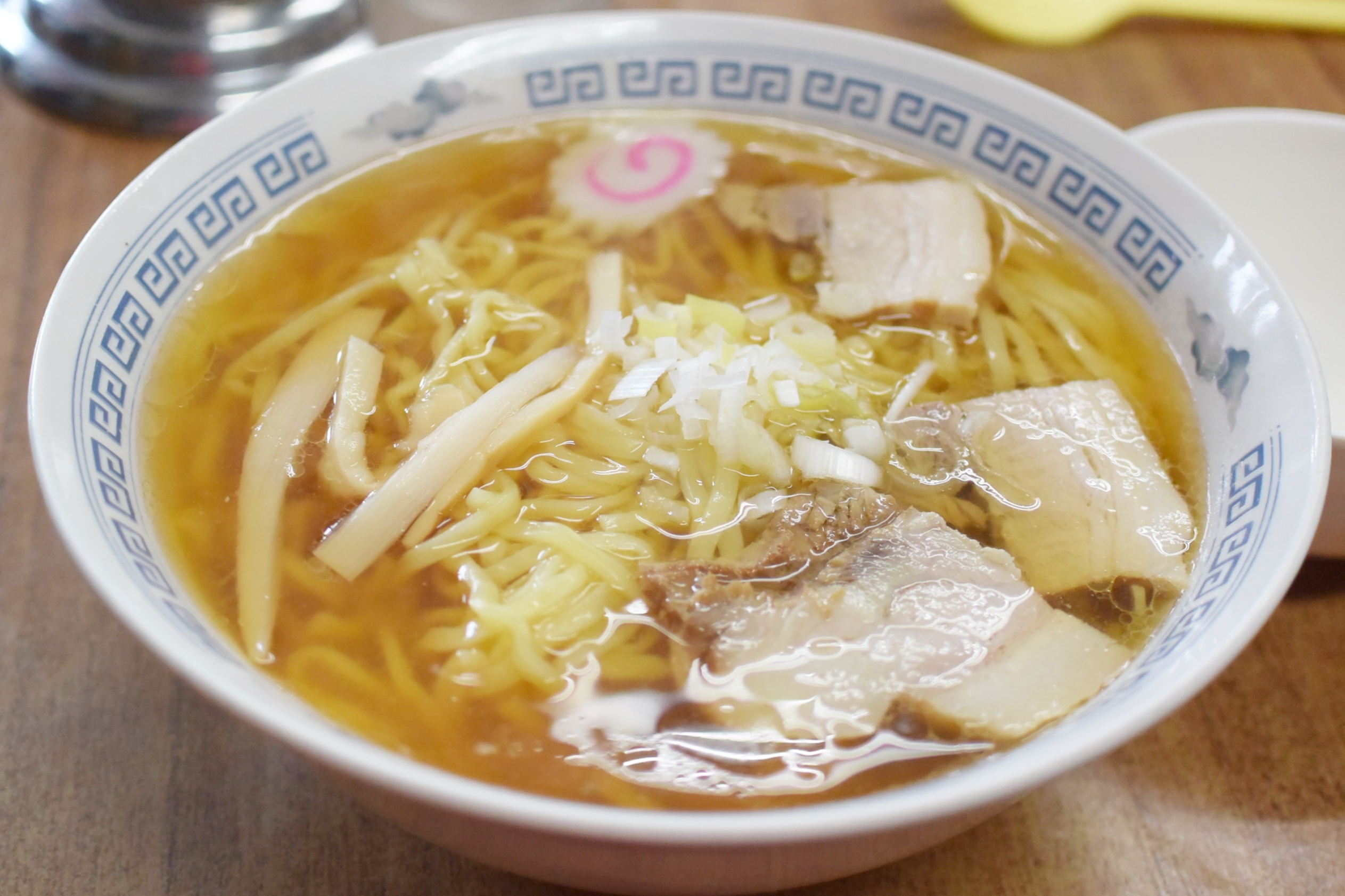 Bowl of ramen with sliced pork, narutomaki fish cake, noodles, and chopped scallions in a soy broth.