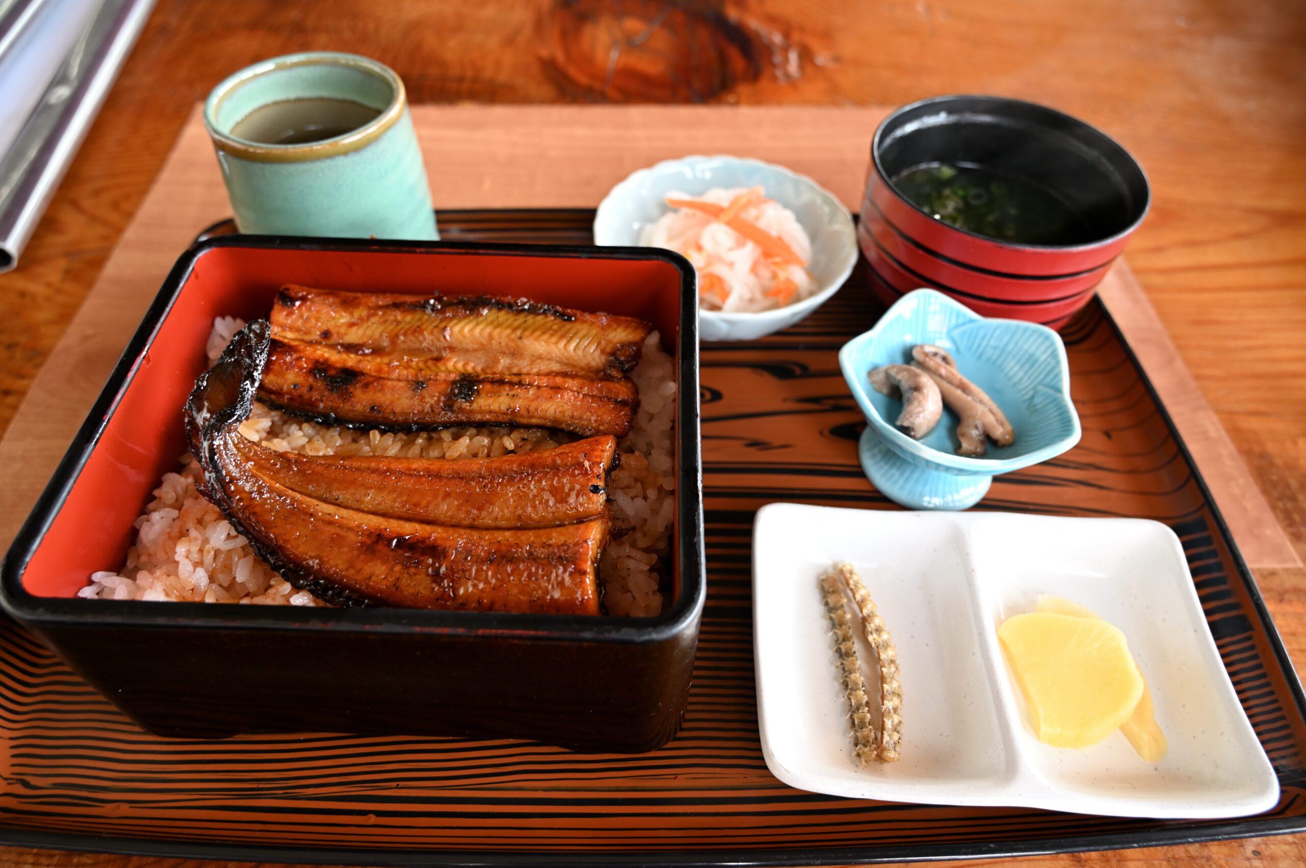 Japanese eel on rice in a lacquered bento box with miso soup, pickles, and side dishes on a wooden tray.